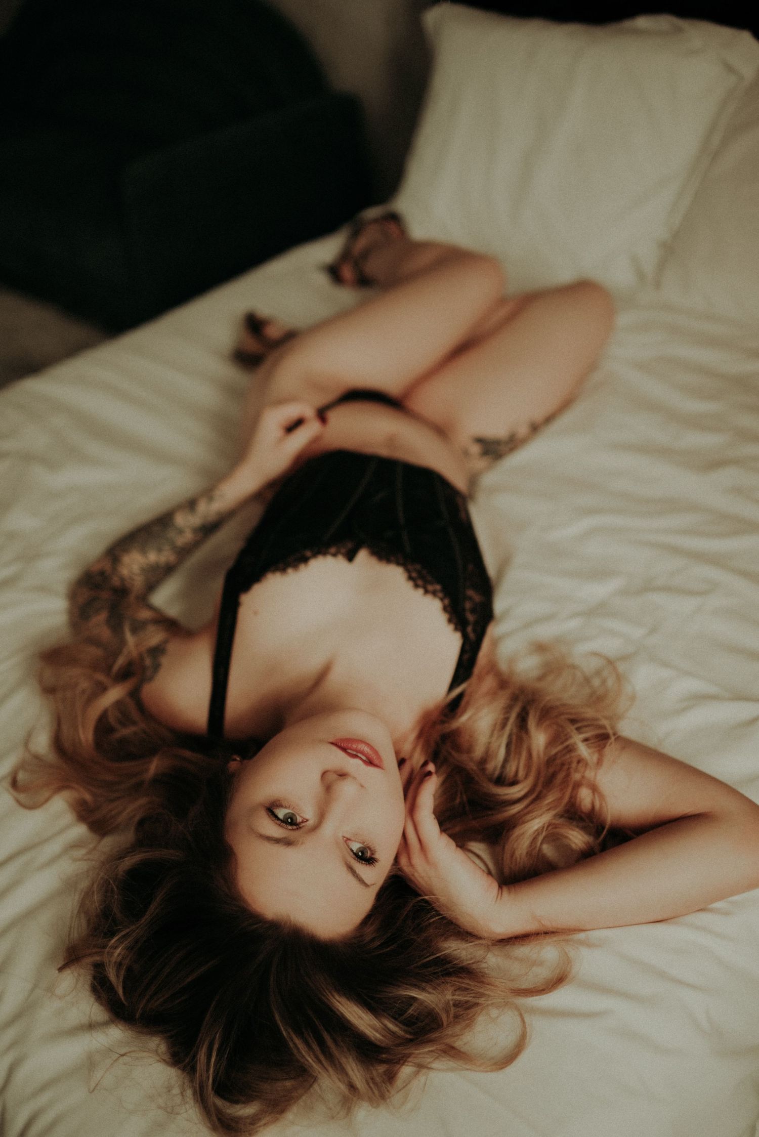 Artistic boudoir photo series showing a person in black lingerie lying gracefully on white bedding in moody lighting.