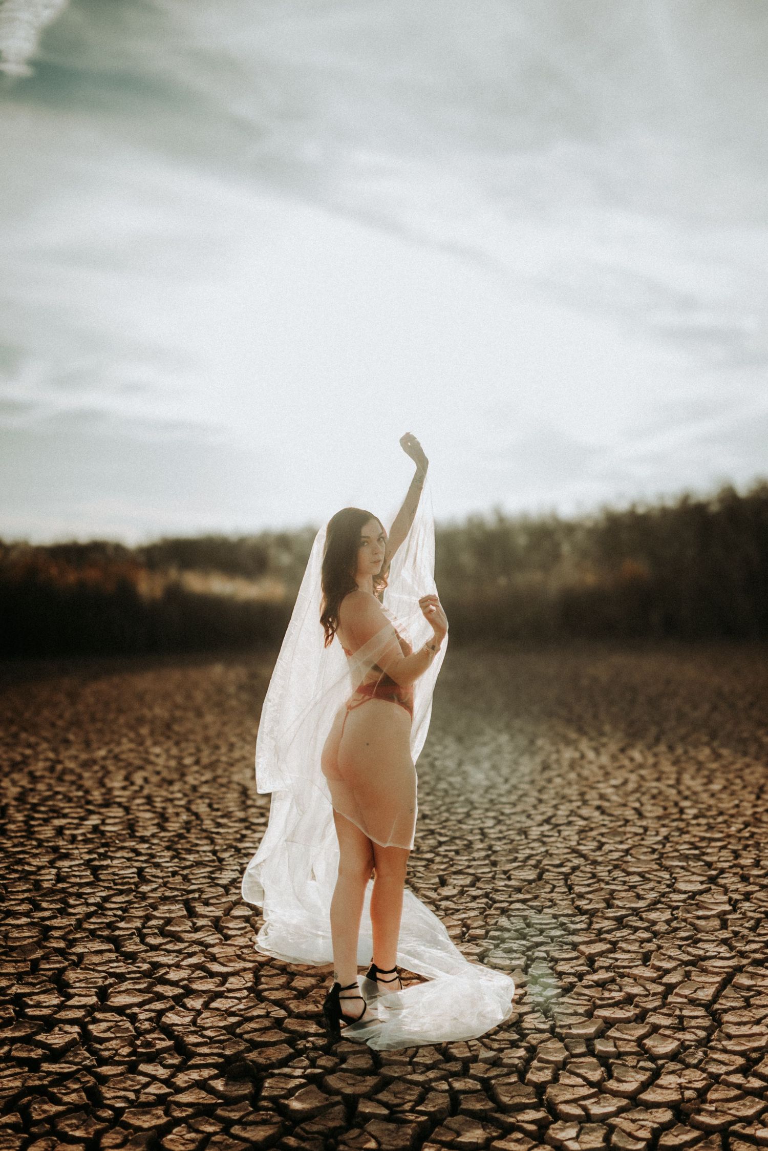 A white wedding veil flows dramatically across cracked desert earth under a moody cloudy sky.