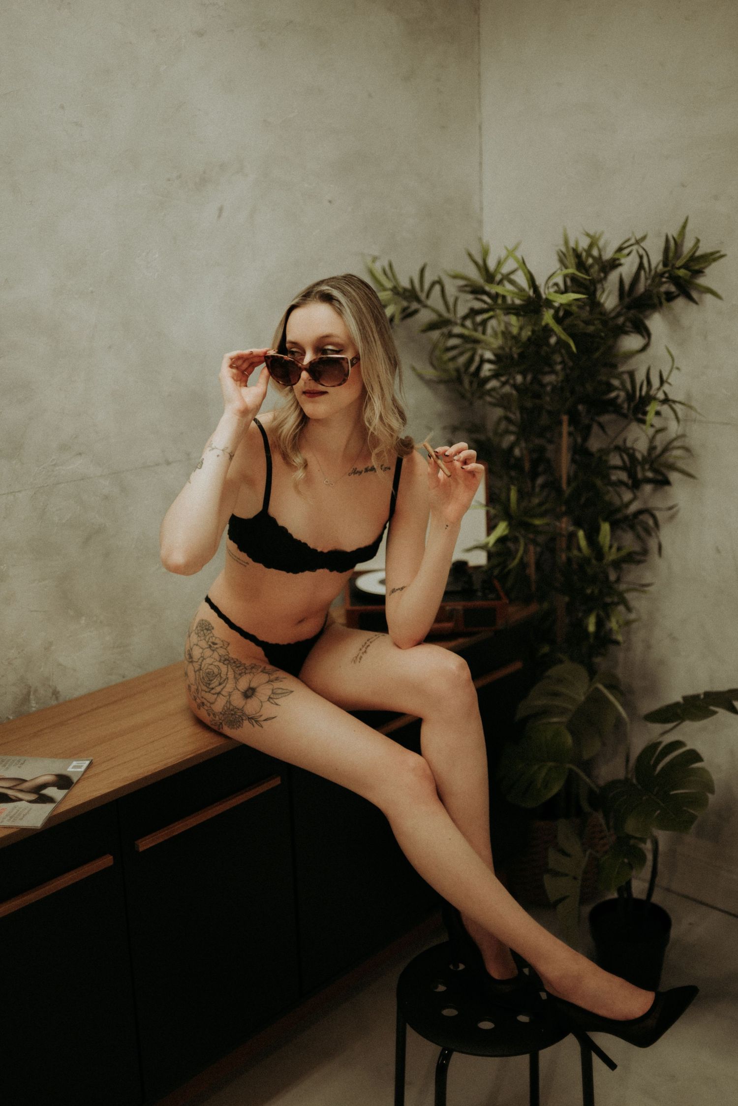 Woman in black lingerie lounges on wooden bench near indoor plants during moody boudoir photoshoot.