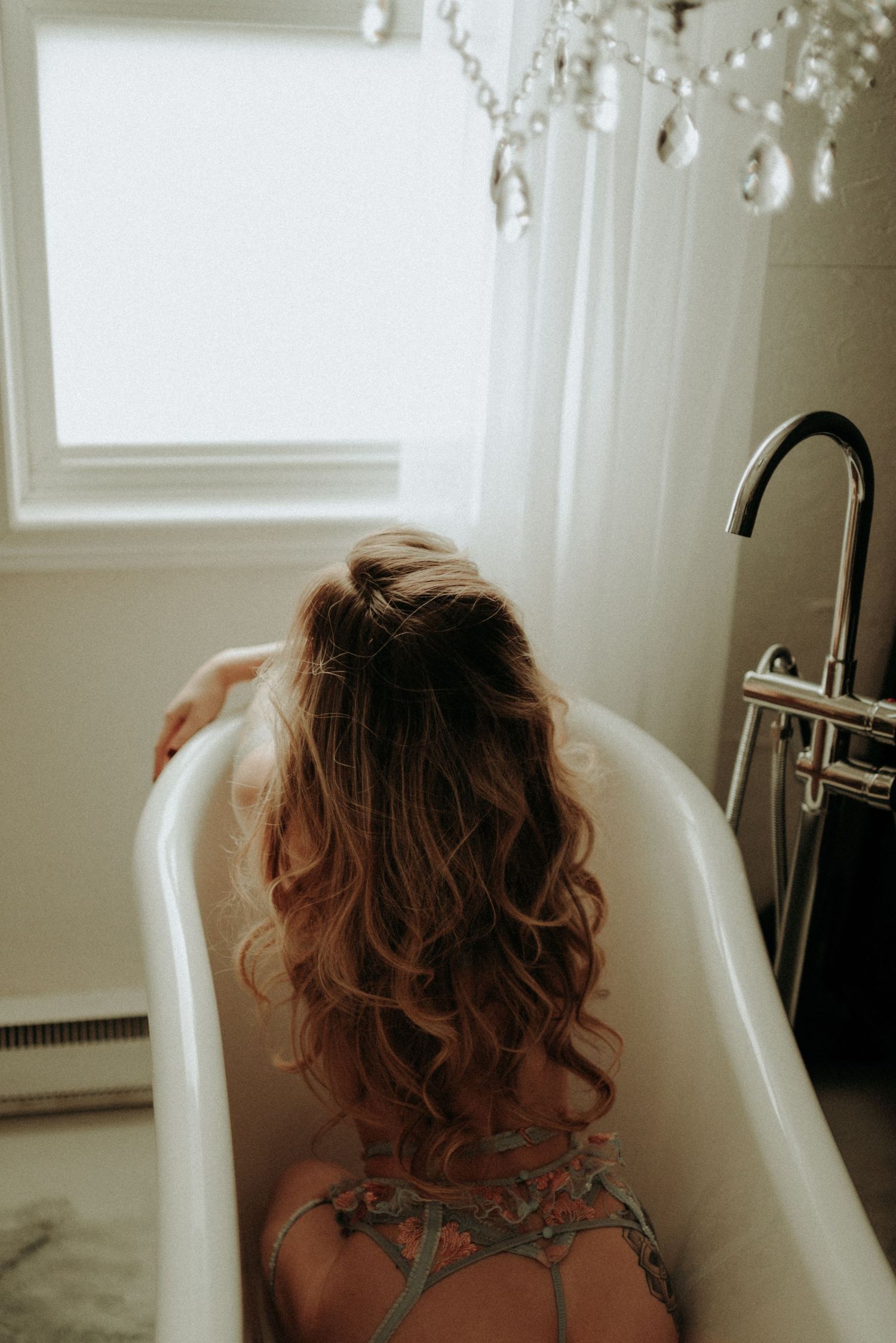 A long-haired person relaxes in a white bathtub under a crystal chandelier, shot from behind.