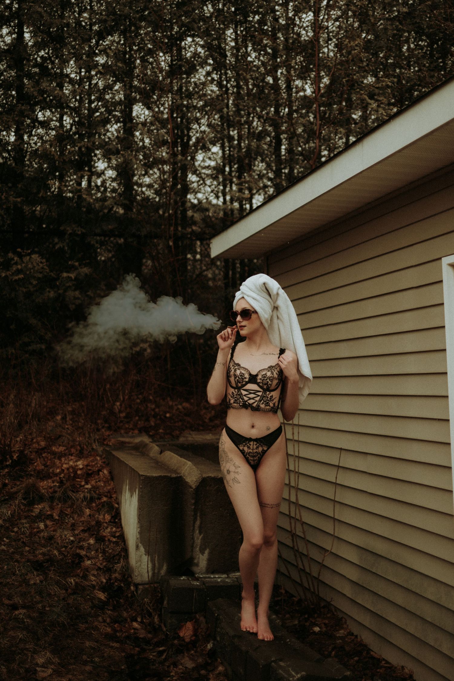 Moody boudoir photo of subject in lingerie smoking against tan house exterior with forest backdrop.