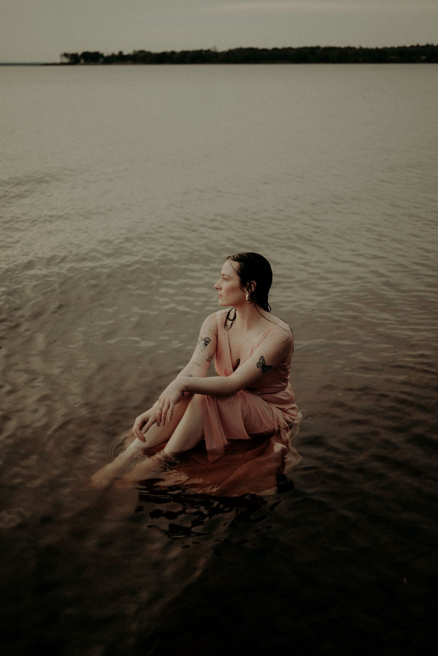A serene water portrait with someone in a rose-colored dress against a cloudy horizon.