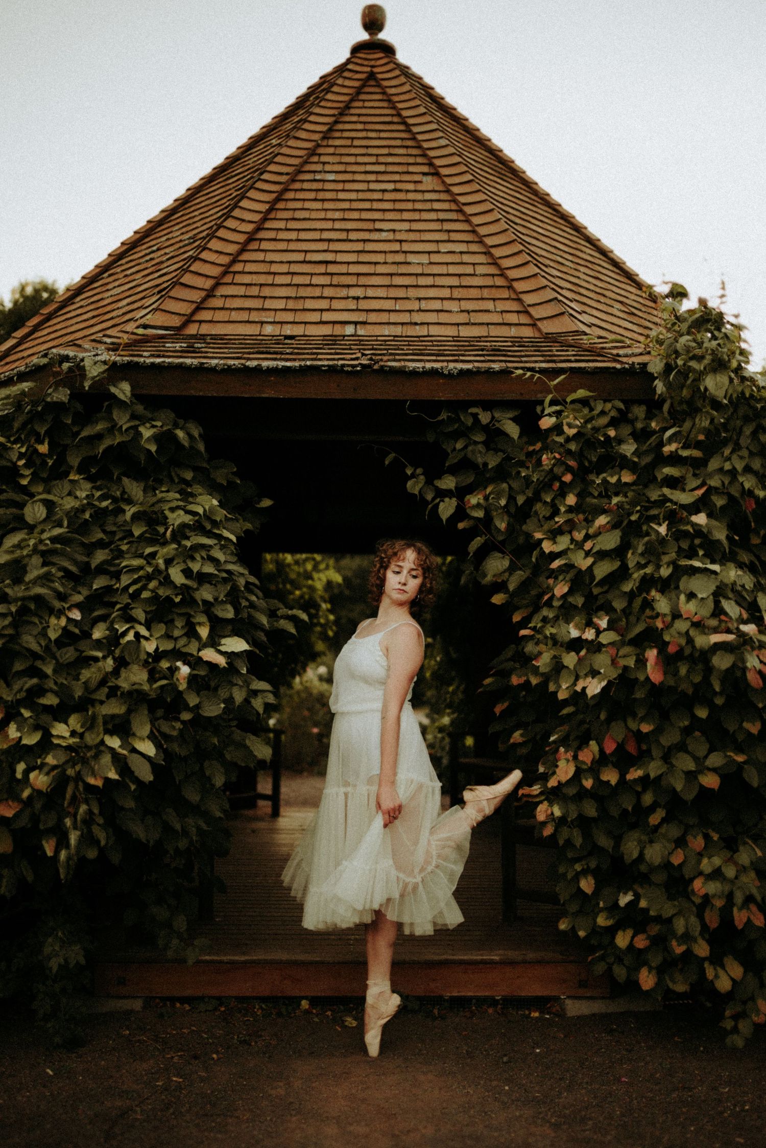 A ballet dancer in a white dress poses en pointe under a wooden gazebo surrounded by lush green vines.
