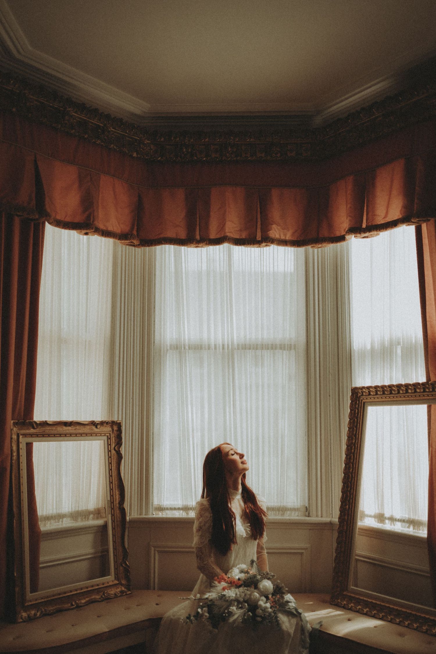 Moody window light illuminates romantic bridal scene with ornate gold frames and russet curtains.