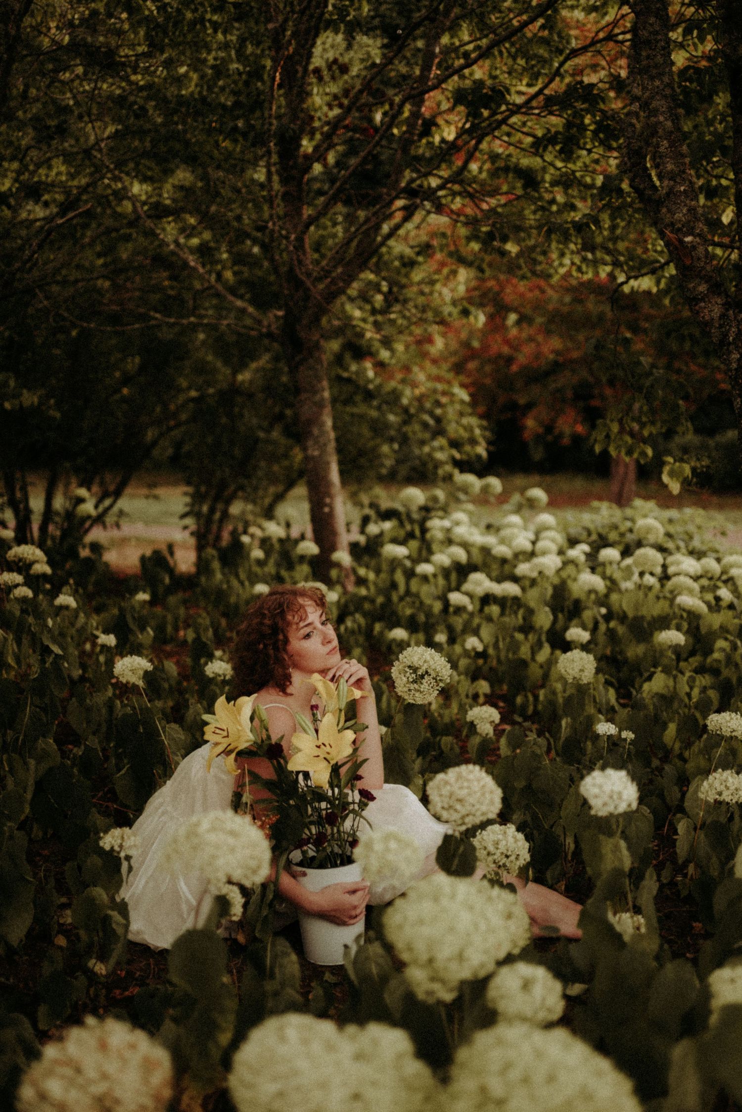 A person in a white dress poses amid a dreamy garden of white peonies and autumn foliage at dusk.