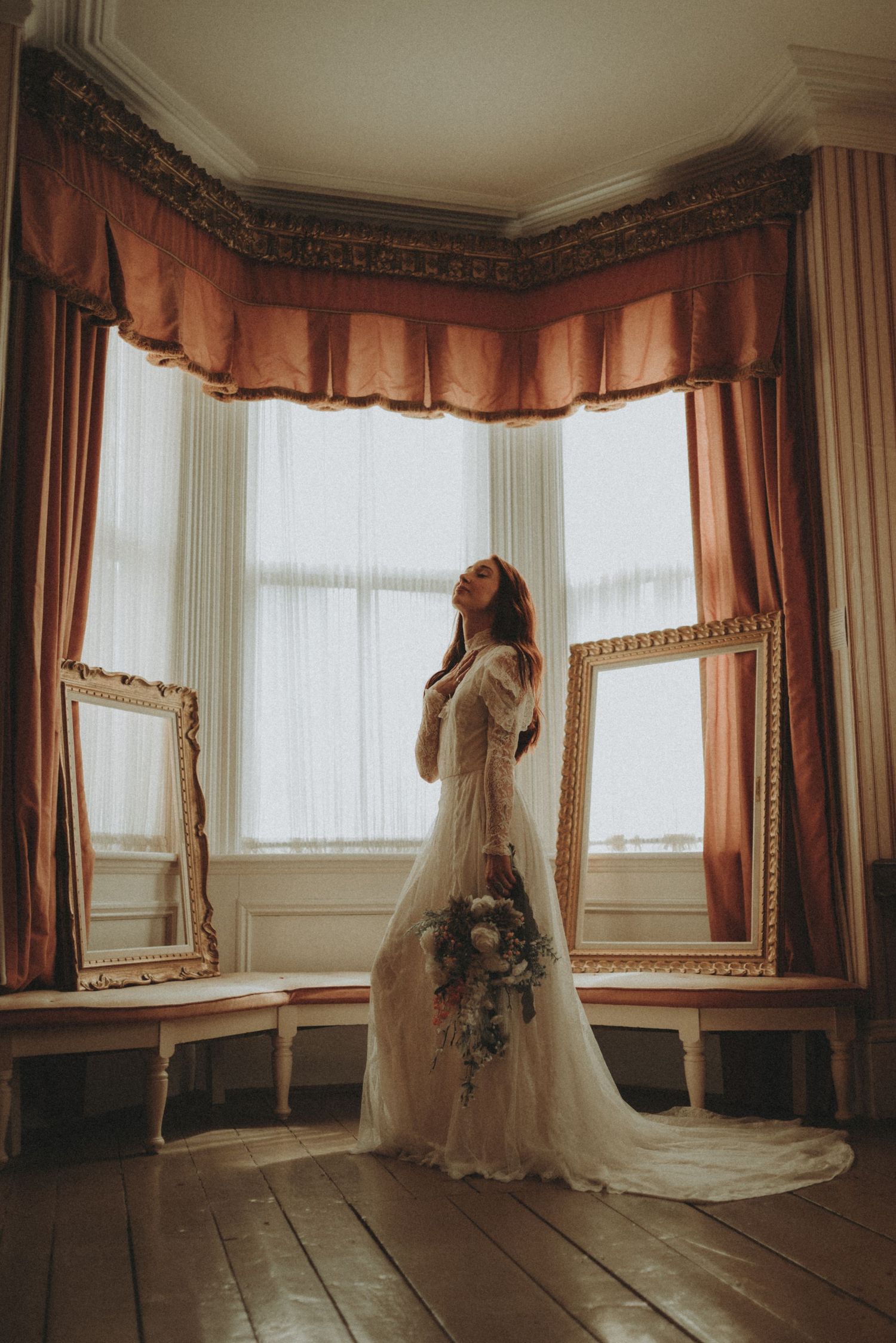 A bride in a vintage lace gown stands gracefully in a bay window alcove with ornate peach curtains and gold-framed mirrors.