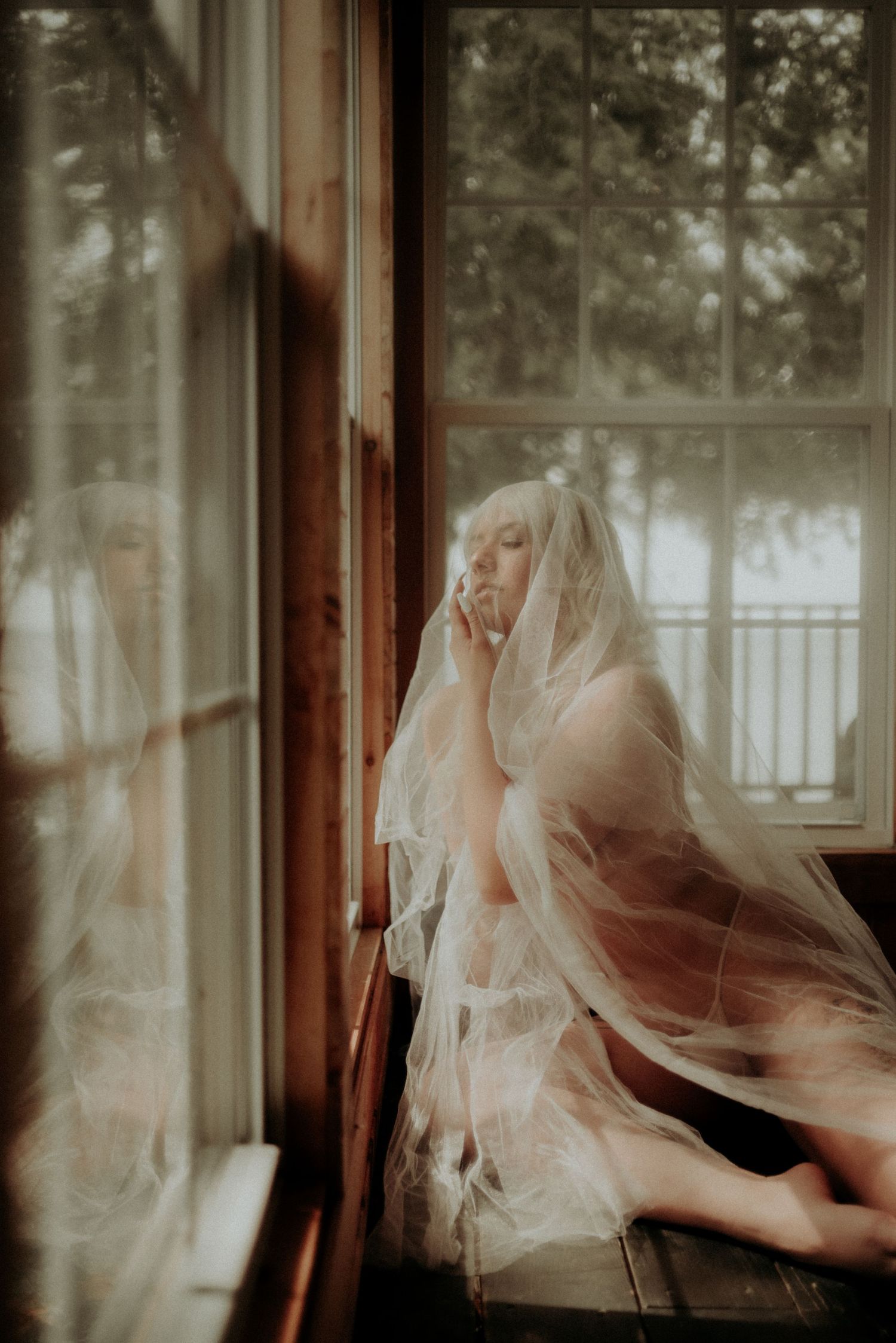 Intimate portrait with sheer veil against window panes in warm sepia tones.