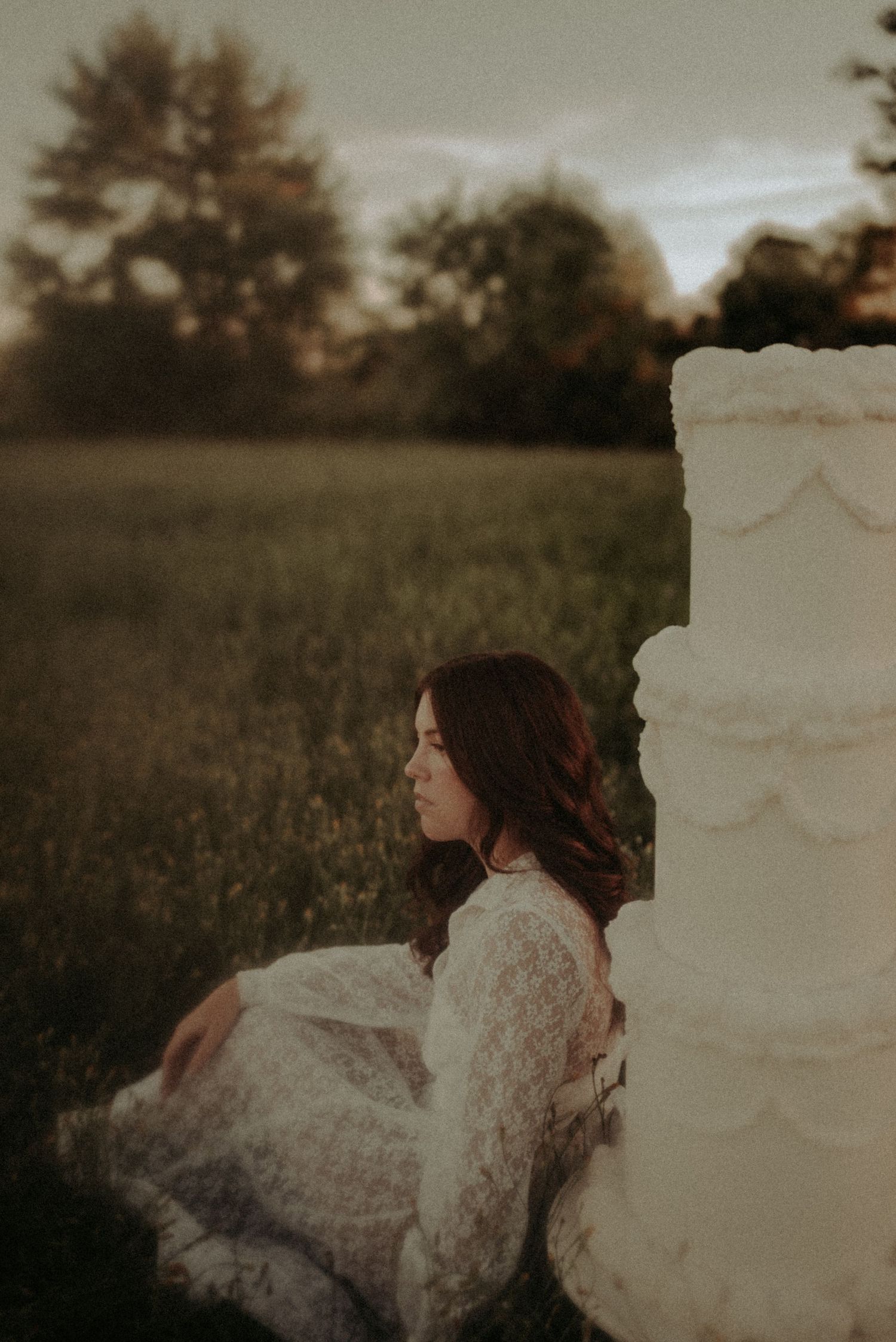 A person in a flowing white lace dress lies on grass at sunset in a dreamy outdoor setting.
