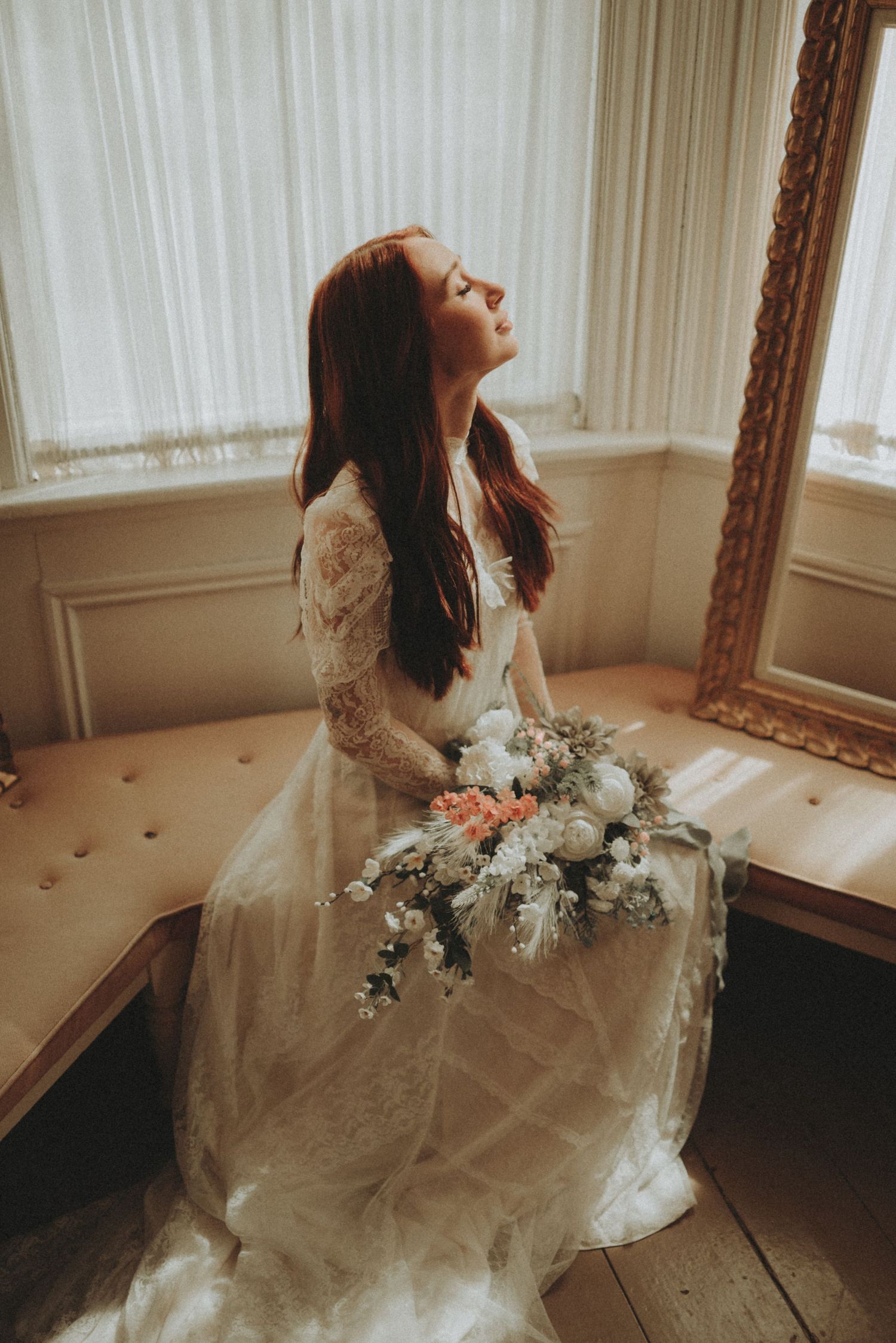 Dreamy vintage bridal portrait in lace gown holding delicate white and blush floral bouquet by sunlit window.