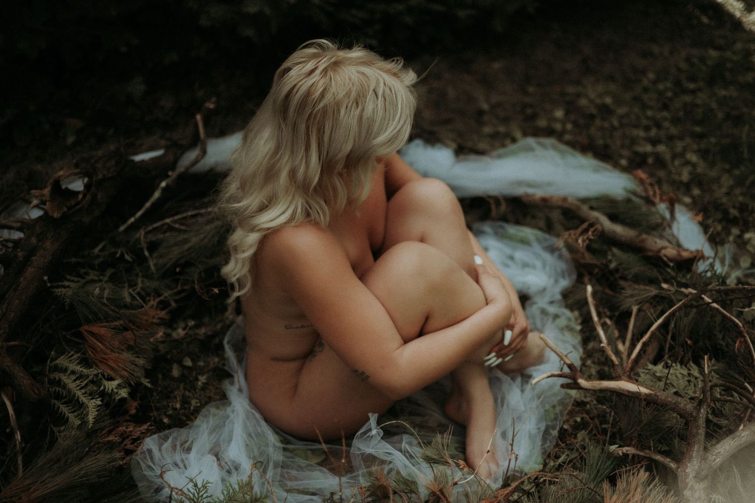 Artistic nude study photographed outdoors with natural lighting and forest elements.