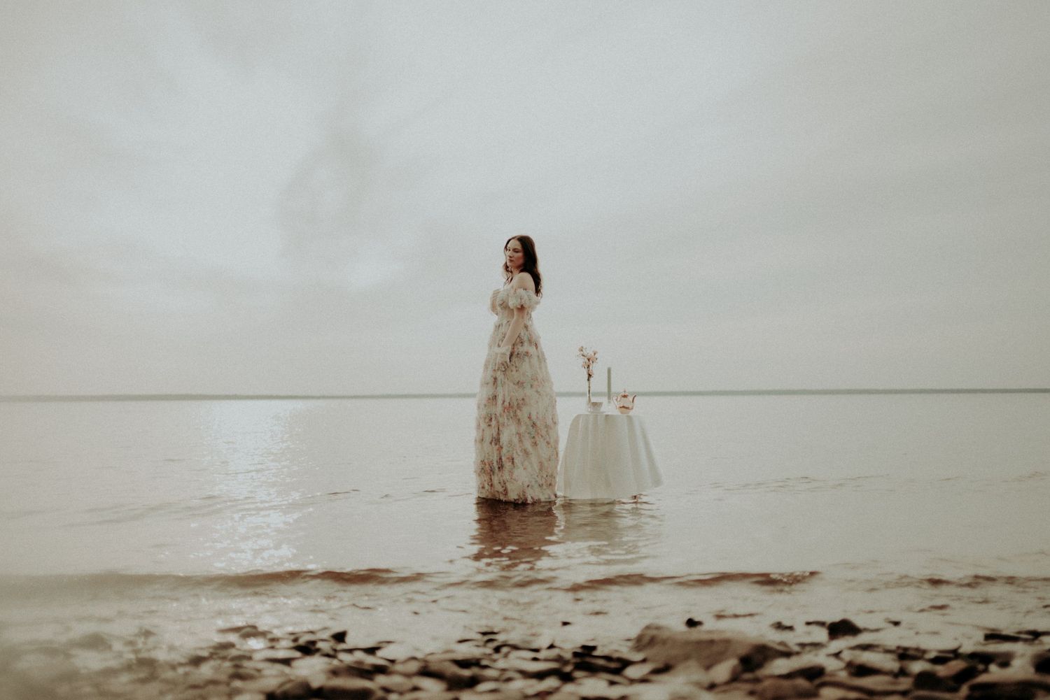 A dreamy figure in floral dress stands in shallow water with white table setting creating ethereal atmosphere.