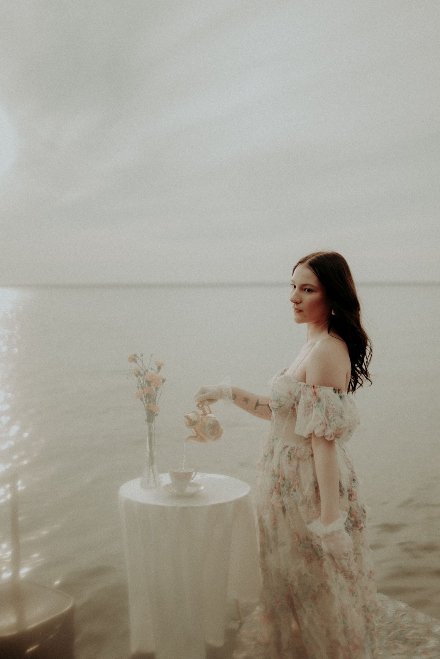 A dreamy photoshoot in shallow water featuring a floral dress and small table with delicate flower arrangement.