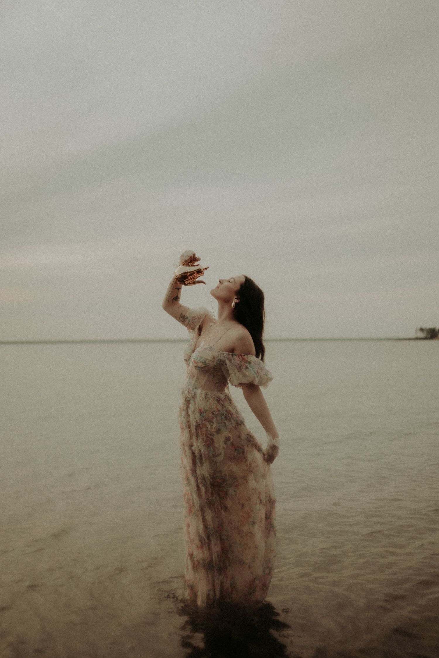 Figure in vintage floral dress poses dramatically in shallow water at sunset.