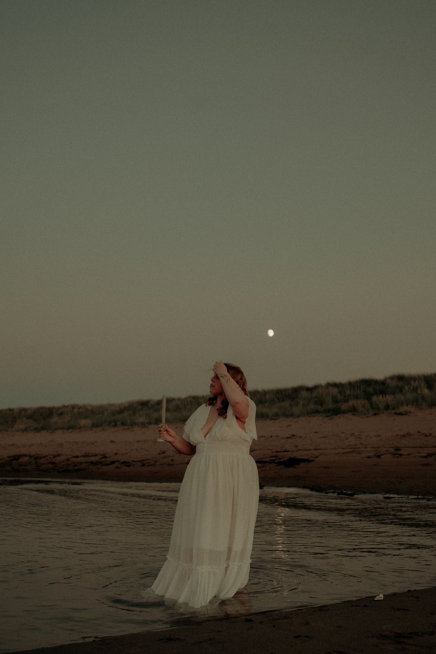 Dramatic beachside portrait with moon visible in muted evening sky and flowing white fabric.
