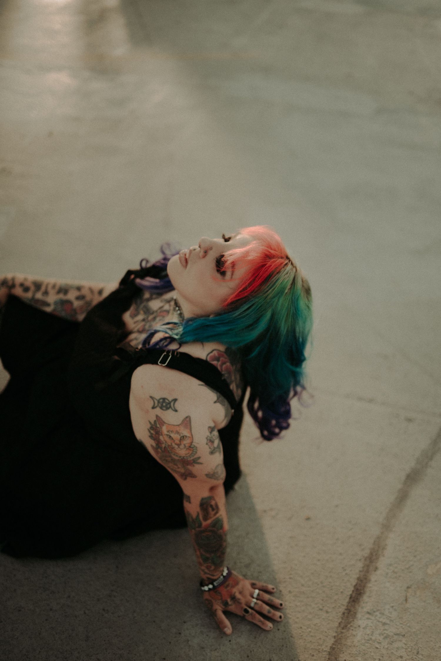 A person with colorful hair and tattoos lies on a concrete floor in an artistic pose.