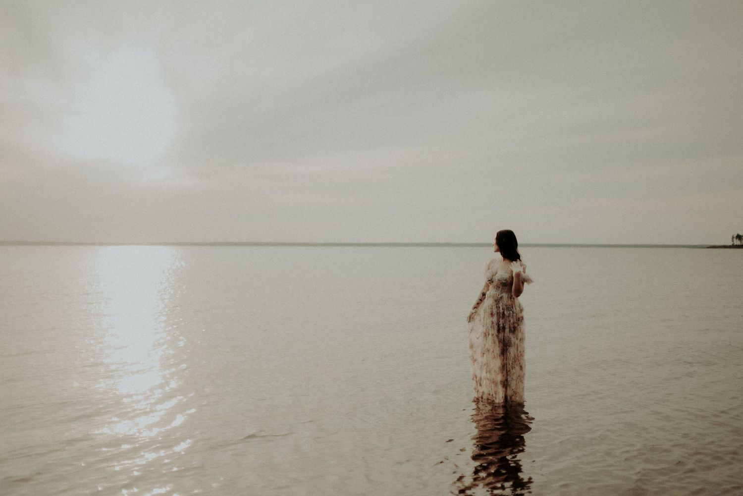 Minimalist landscape photo with figure in floral dress standing in still waters at dusk.