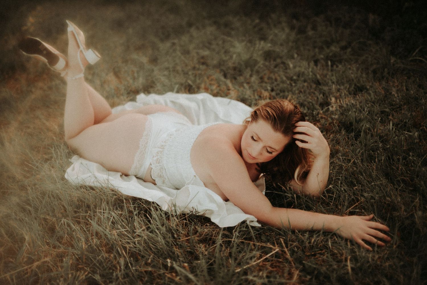 A figure in a flowing white dress lies gracefully on dark grass in a dreamy outdoor setting.