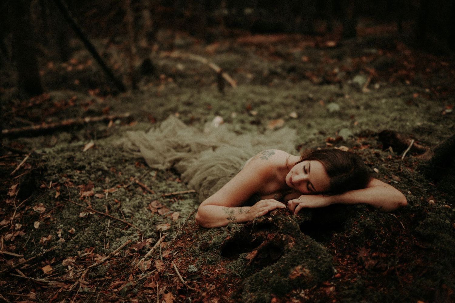 A person lies on autumn leaves in a dark forest setting creating a moody outdoor portrait.