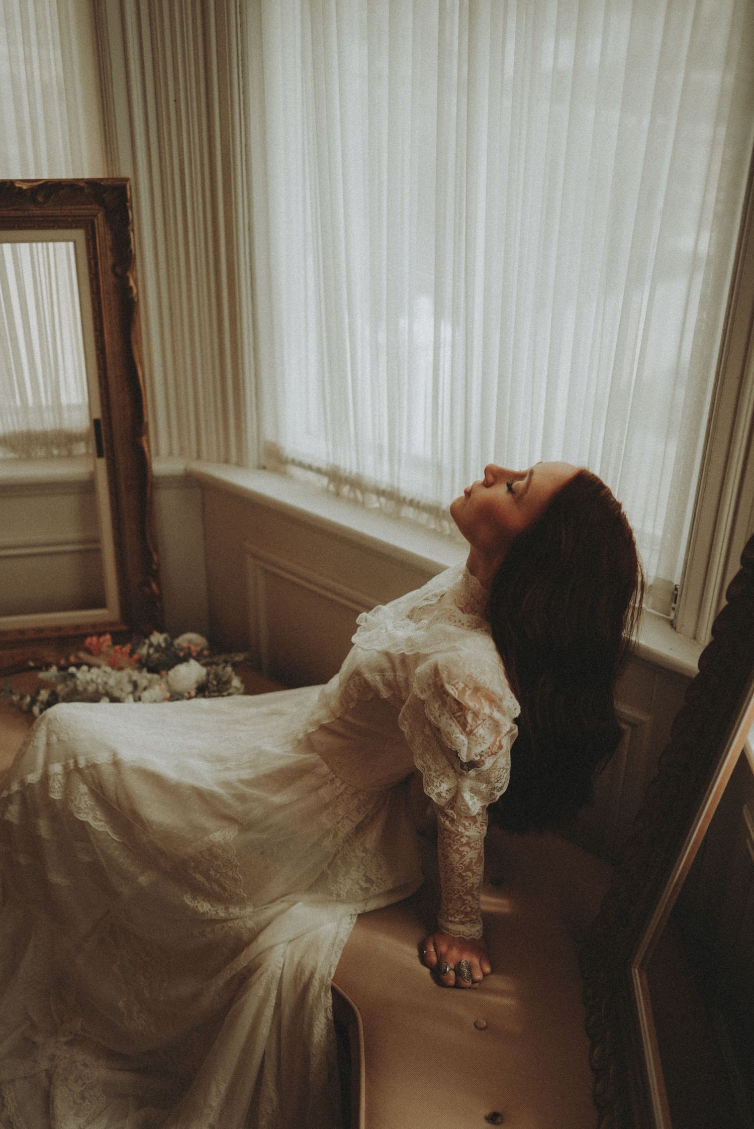 Moody vintage-style bridal photo with flowing lace dress in natural window light against white curtains.
