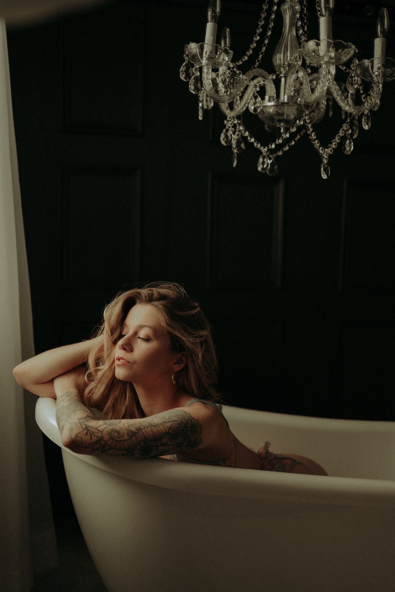 A moody boudoir photo shows a bathtub scene with crystal chandelier and dramatic shadows.