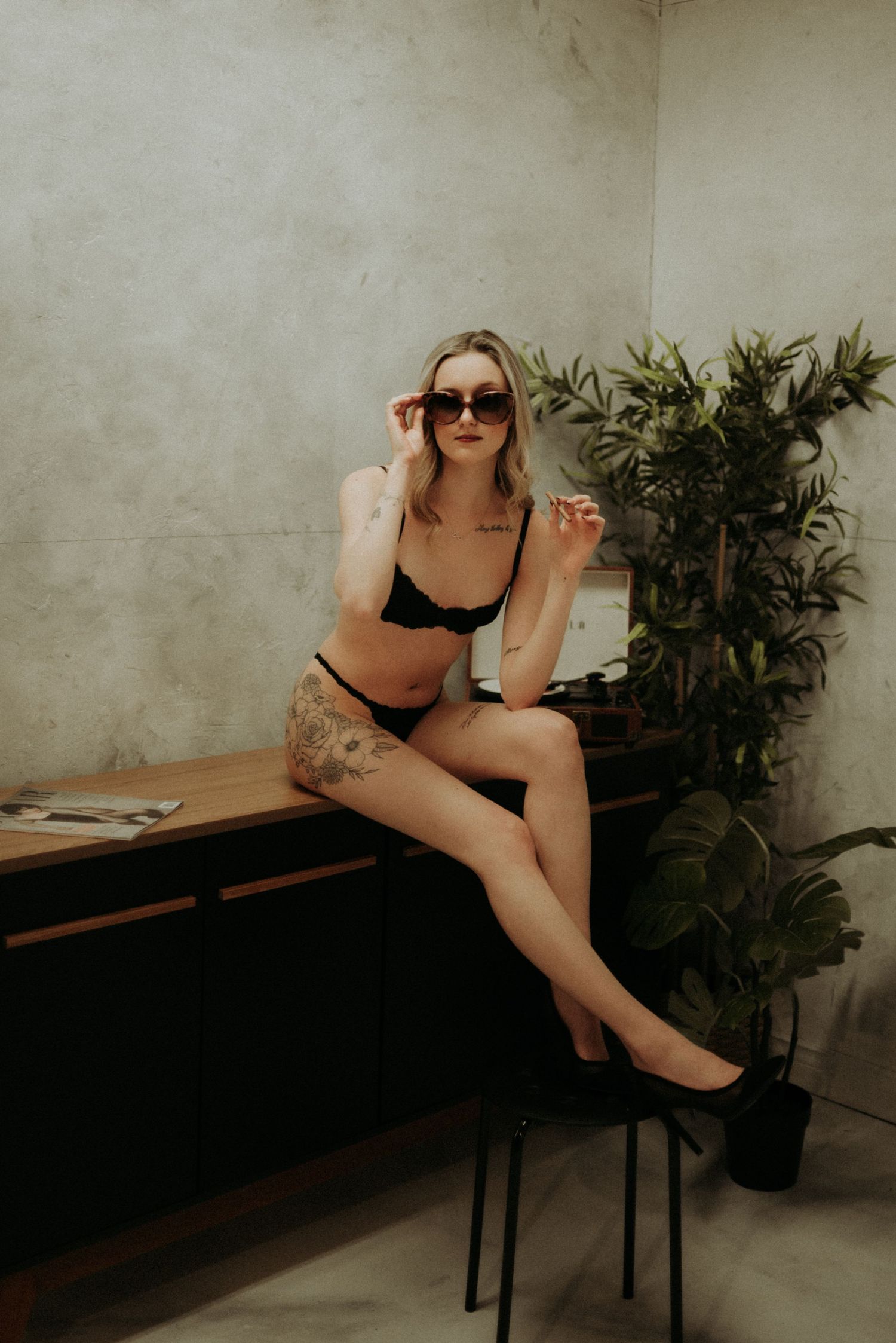 A person in black lingerie poses on a wooden bench against a concrete wall with indoor plants in the background.