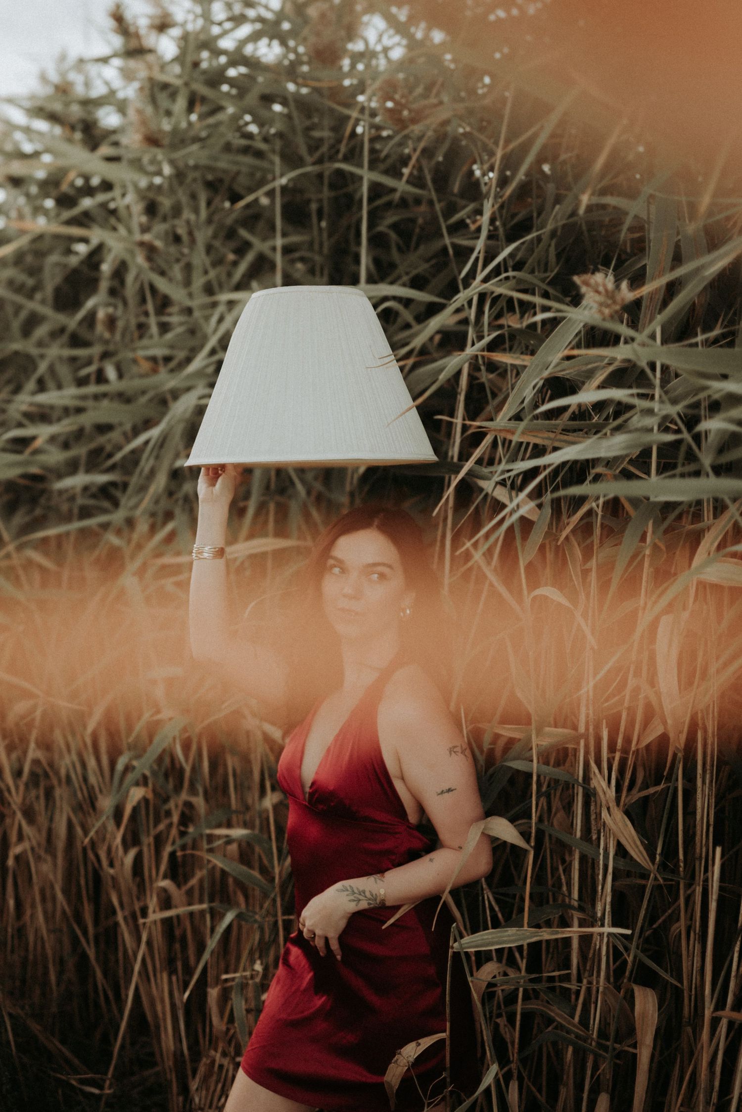 A person in a burgundy dress poses in tall grass with a white parasol at sunset creating a dreamy atmosphere.