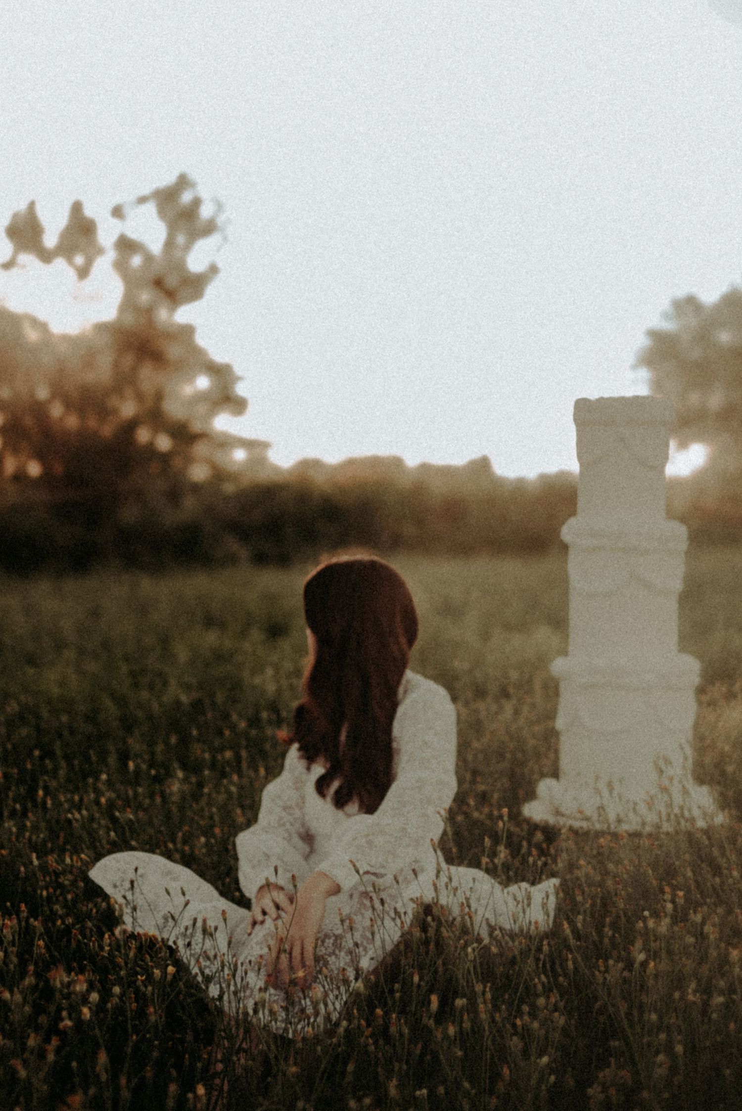 A person in a white dress sits in a field at sunset with a decorative white pillar in the background.