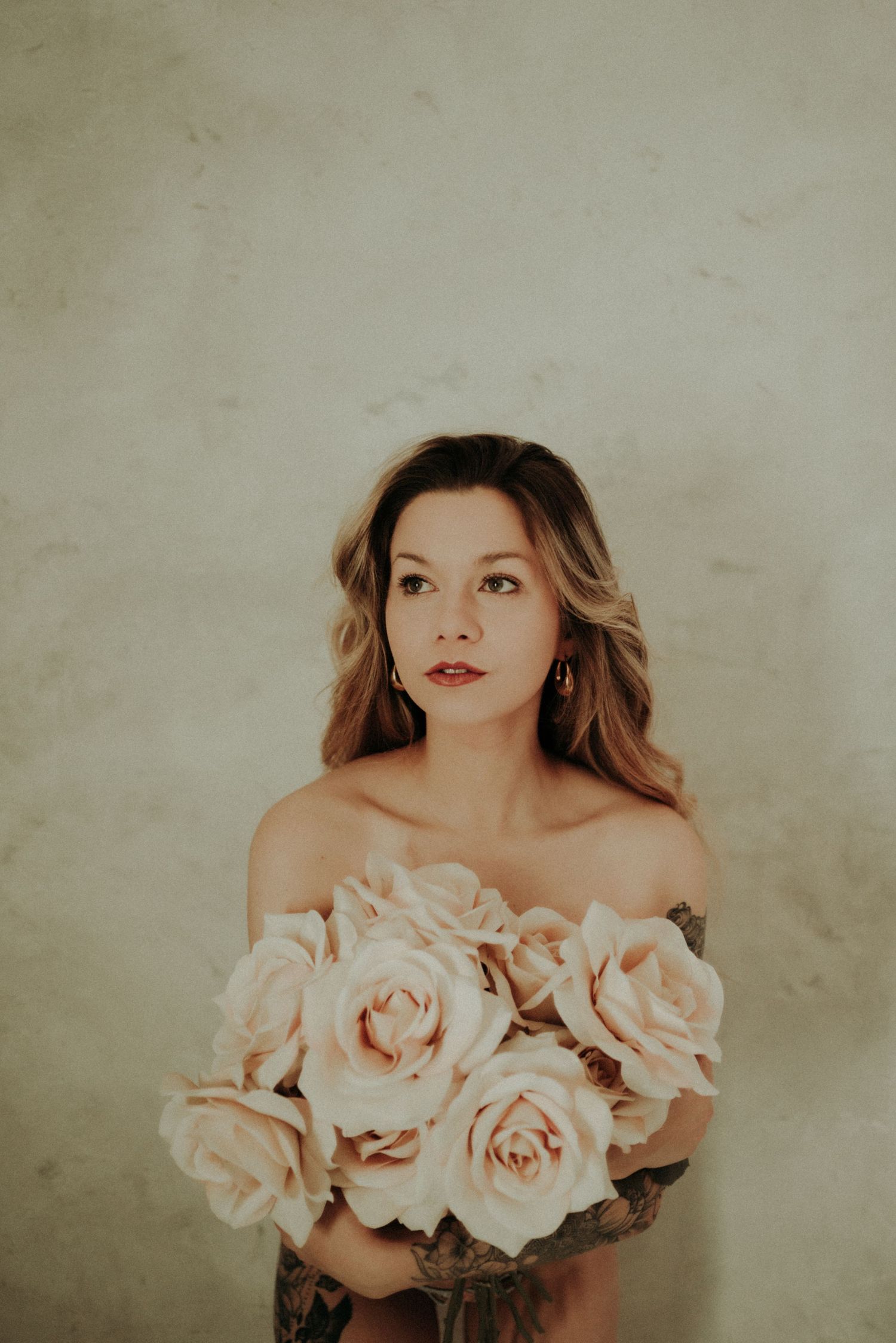 Ethereal portrait with pale roses against beige background in delicate off-shoulder pose.