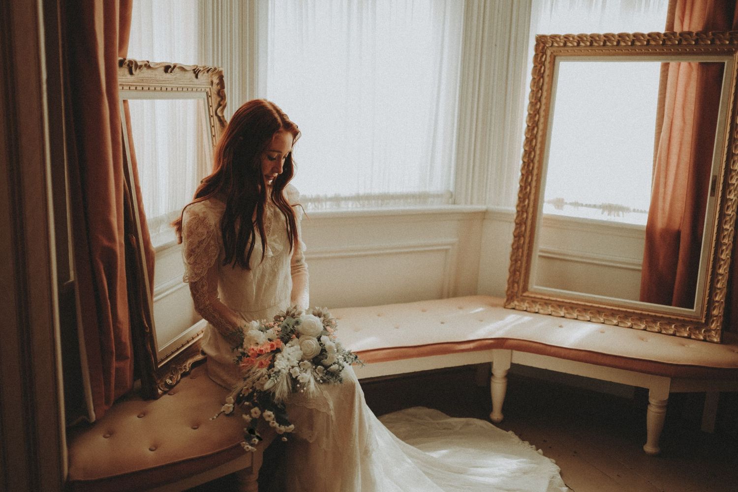 A bride in a white gown sits by a window holding a delicate floral bouquet in a vintage-style room with ornate mirror.