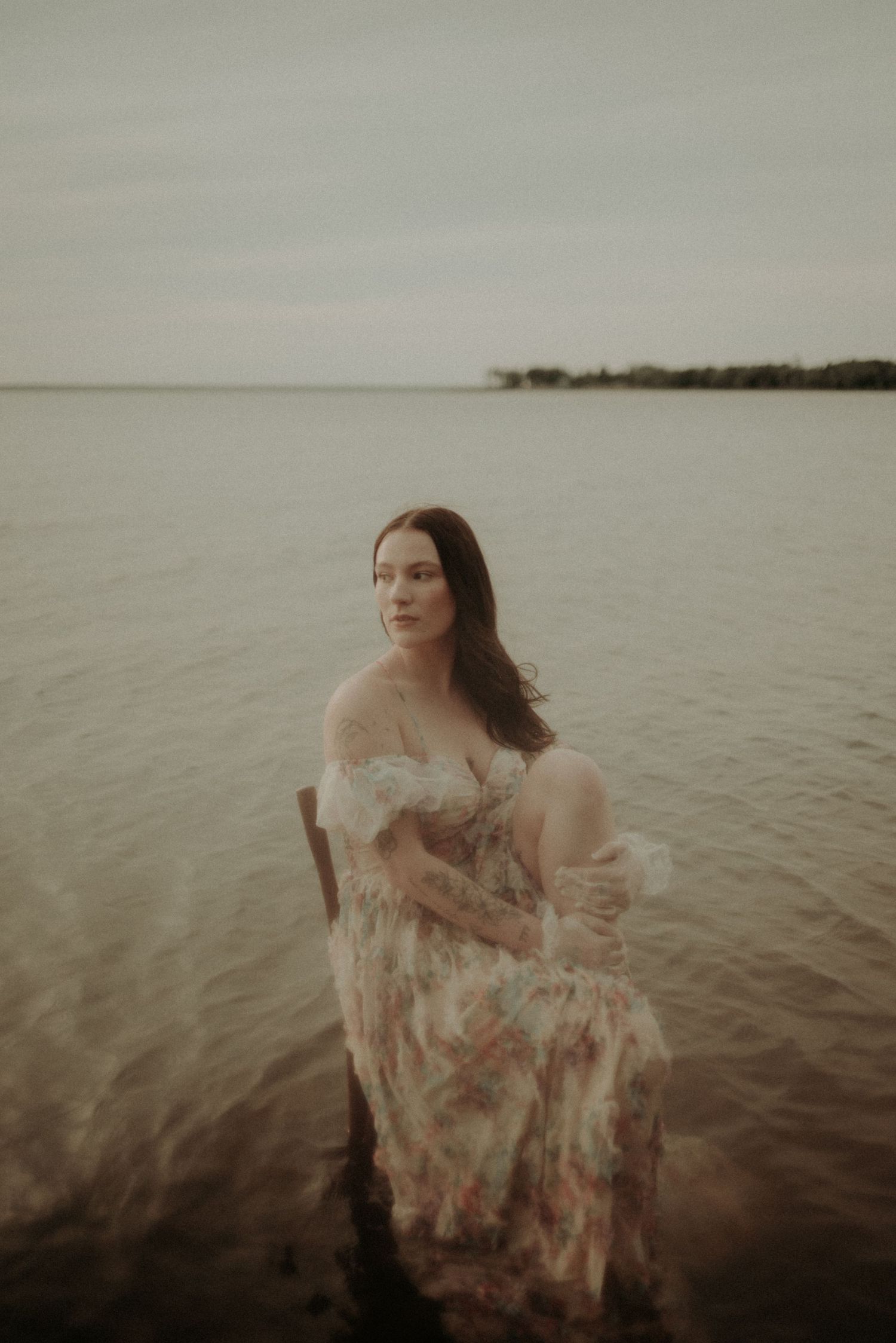 A dreamy portrait in shallow water wearing a flowing lace dress against a misty waterfront landscape.