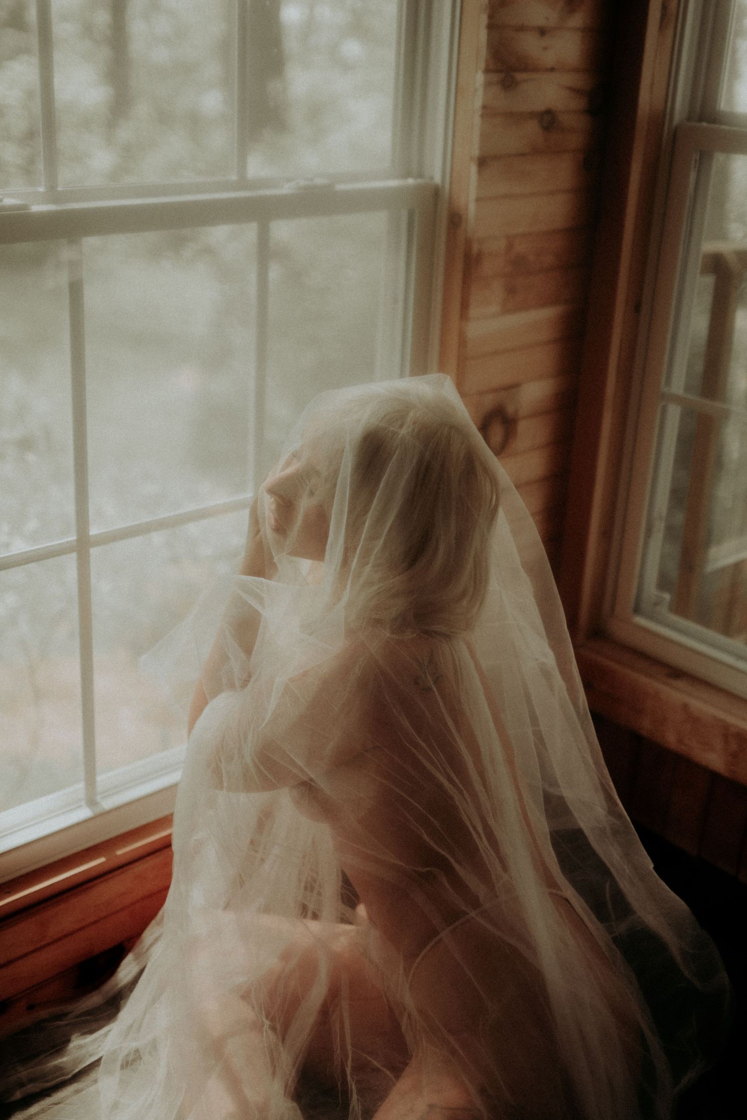 Ethereal figure draped in delicate tulle veil poses by window with soft natural lighting.