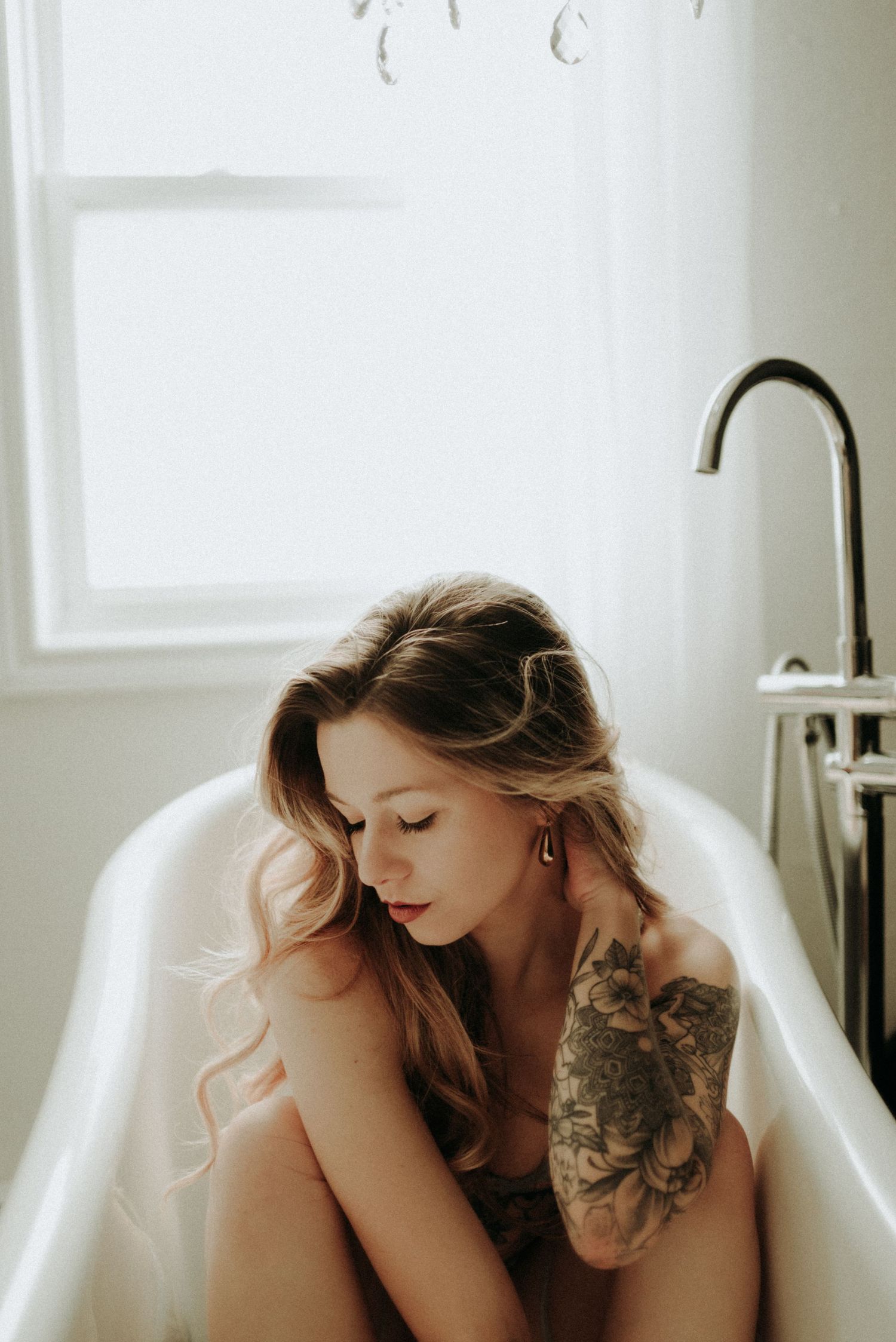 Intimate portrait in white bathroom featuring delicate pose and visible floral tattoo artwork.