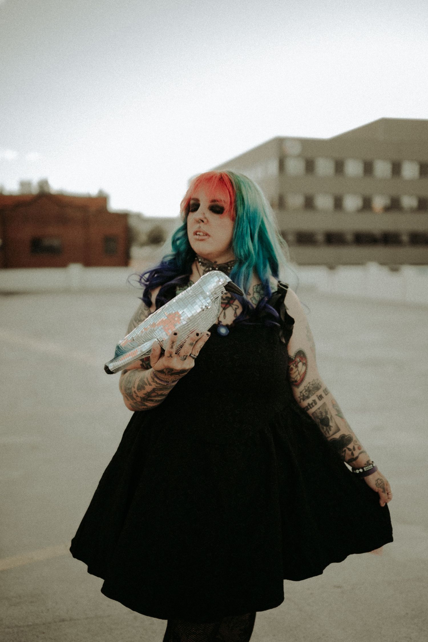 A figure in a flowing black dress and teal hair poses dramatically against an overcast urban backdrop.