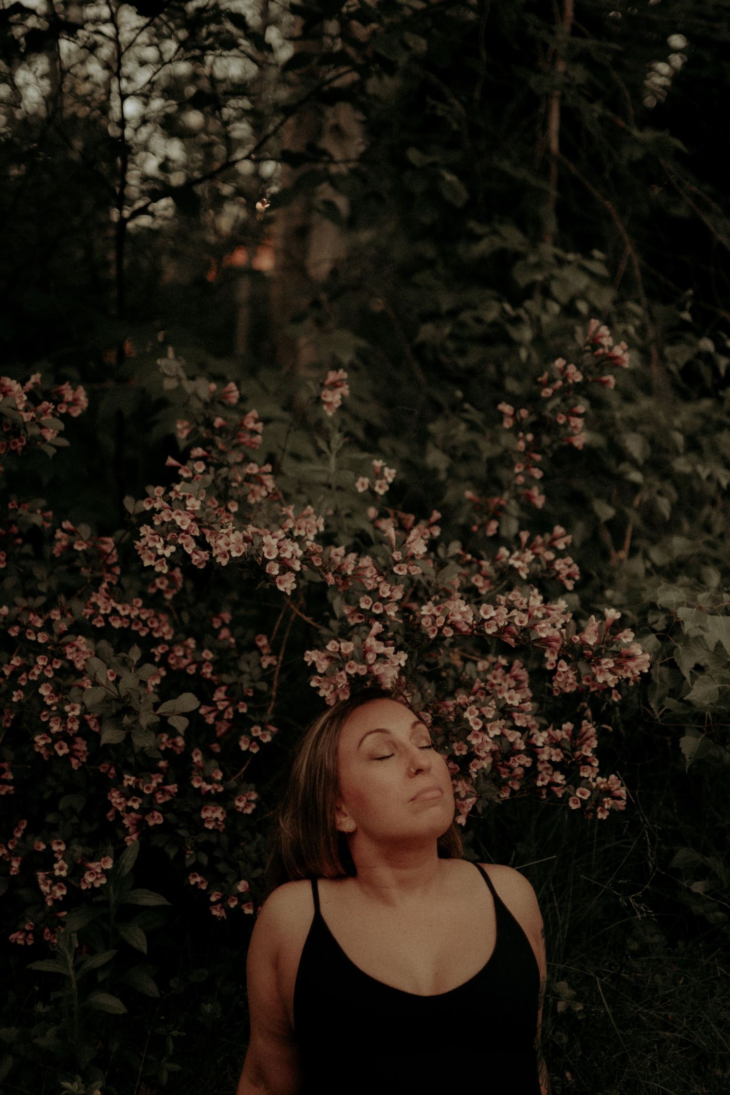 Artistic portrait against pink flowering bush with moody lighting and dark tones.