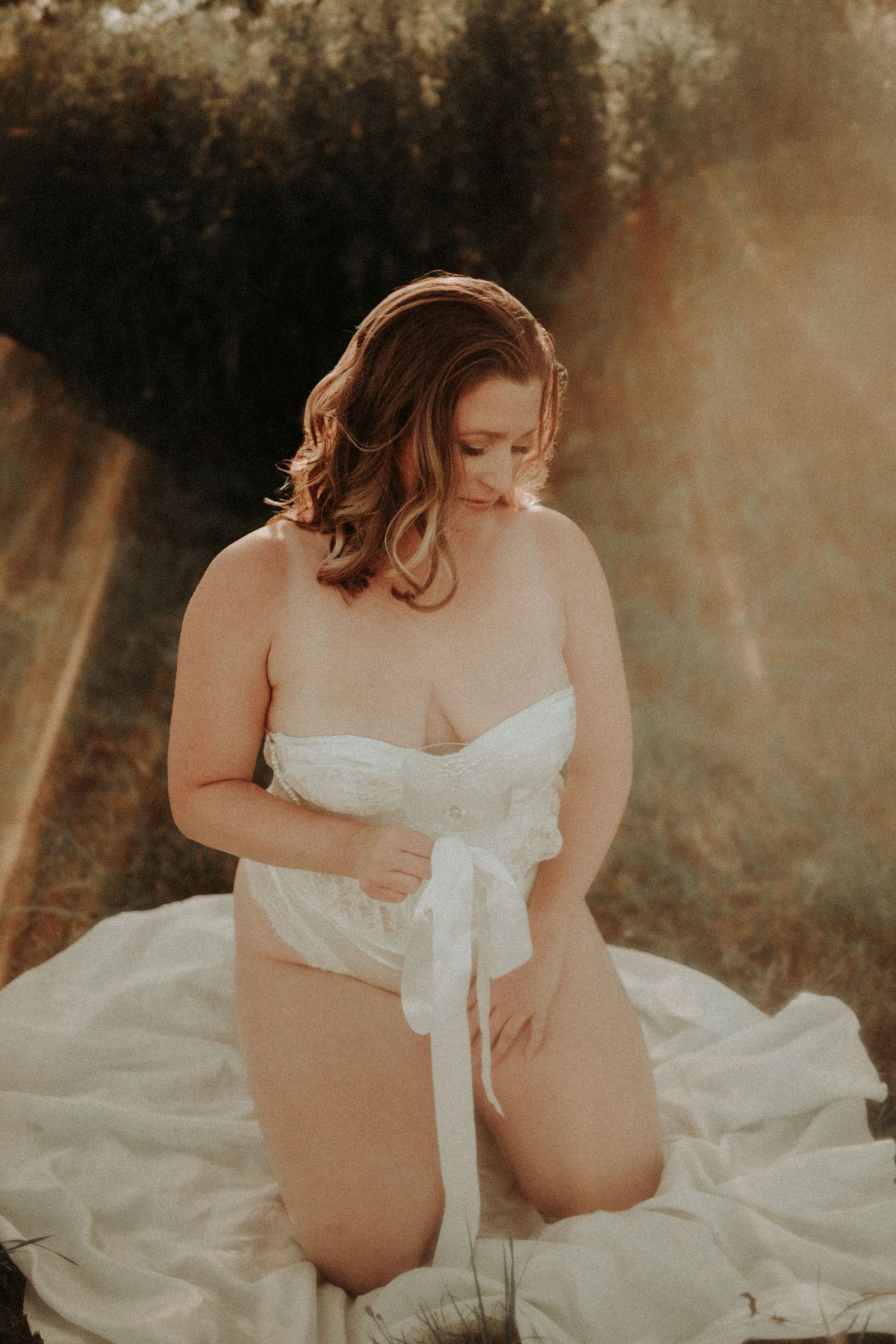 A maternity photoshoot in soft golden light shows a woman in a flowing white dress sitting on the ground.