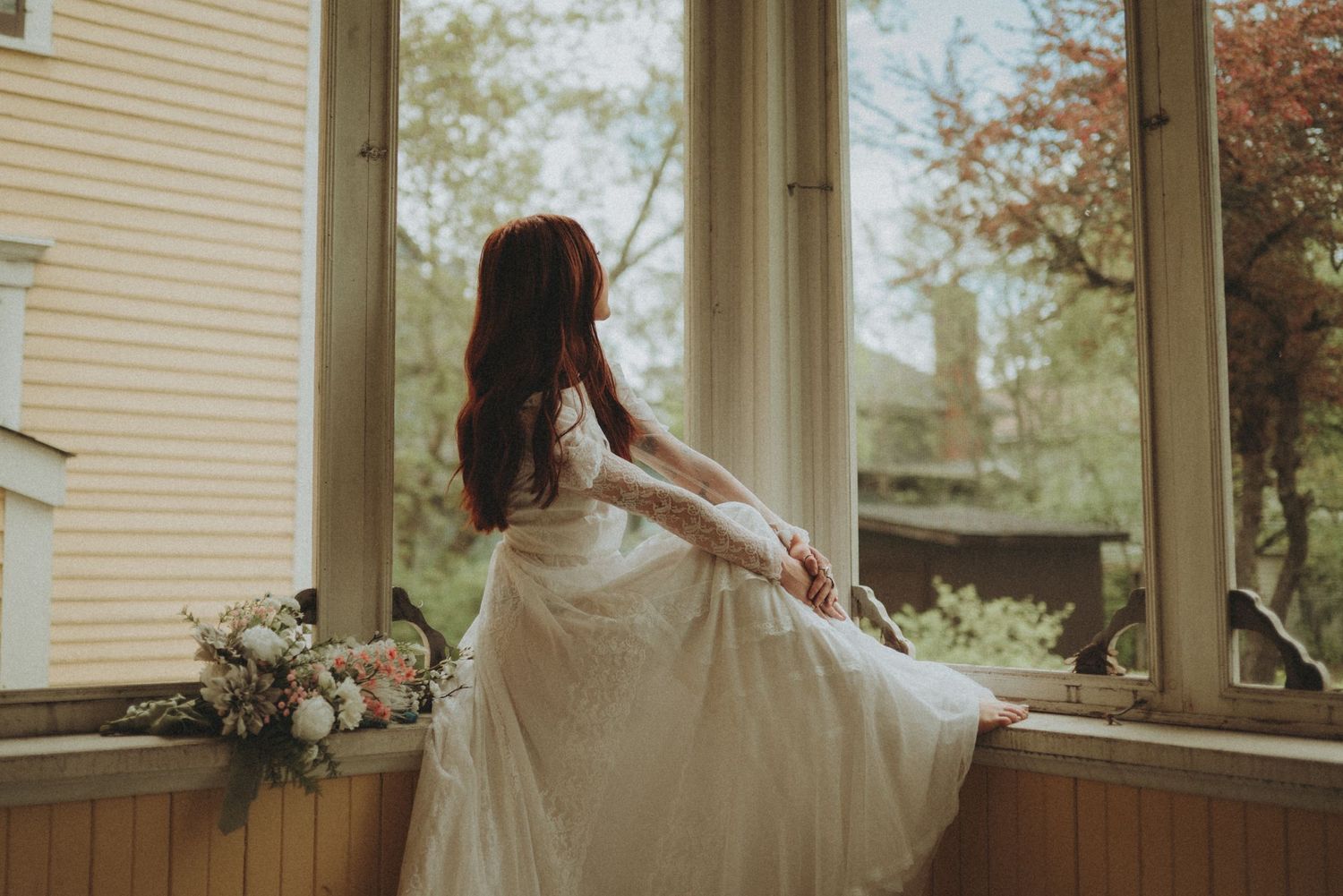 A person in a flowing white dress sits by a sunlit window with a floral bouquet in a vintage-style interior setting.