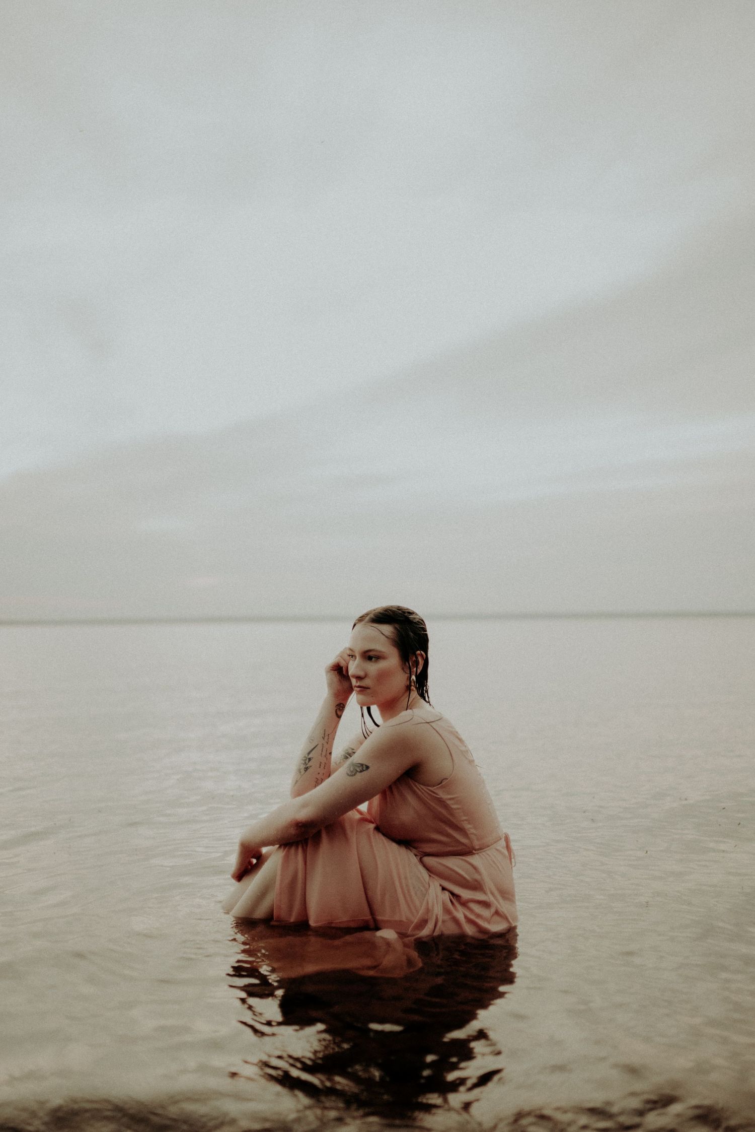 A contemplative figure in pink dress sits in shallow water during golden hour.