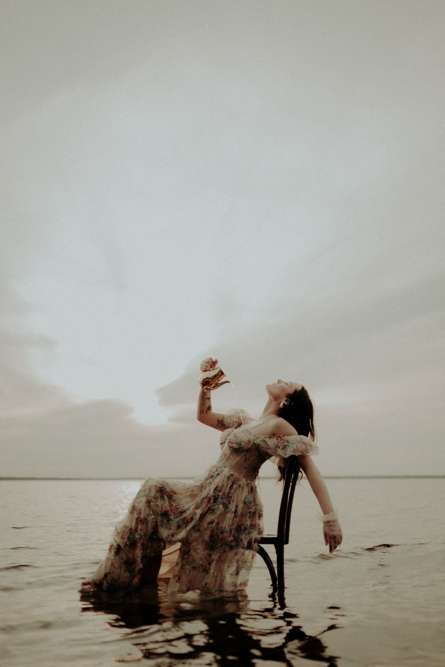 Moody fine art photograph of a person in a floral dress posed on a chair in tranquil waters.