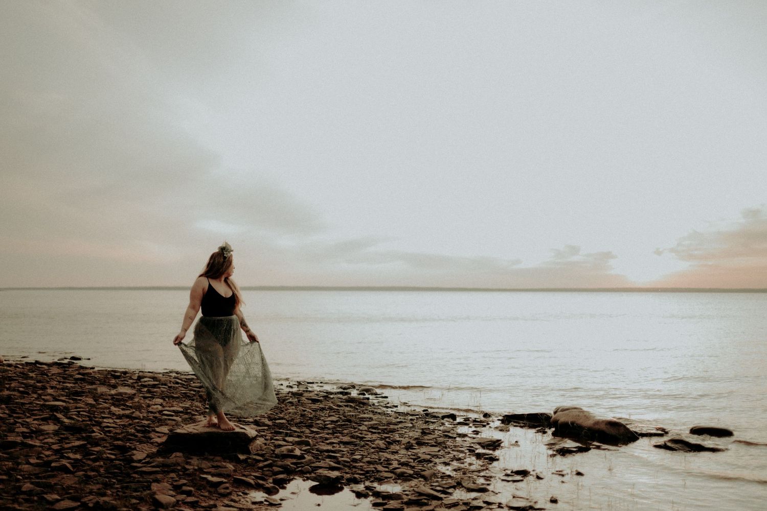 Silhouetted figure in a dress stands on rocky beach at sunset with calm ocean waters in background.