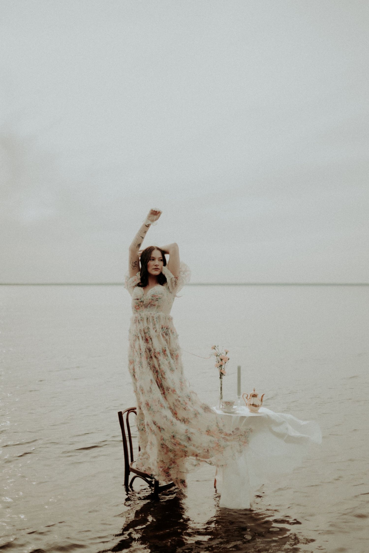Artistic water portrait with flowing dress and vintage table setup against a minimalist waterscape.