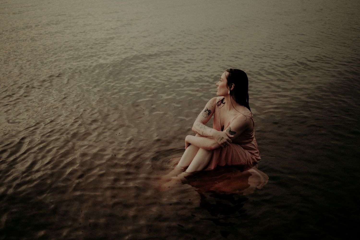 Atmospheric photo of someone in a dusty rose dress sitting contemplatively in dark waters.
