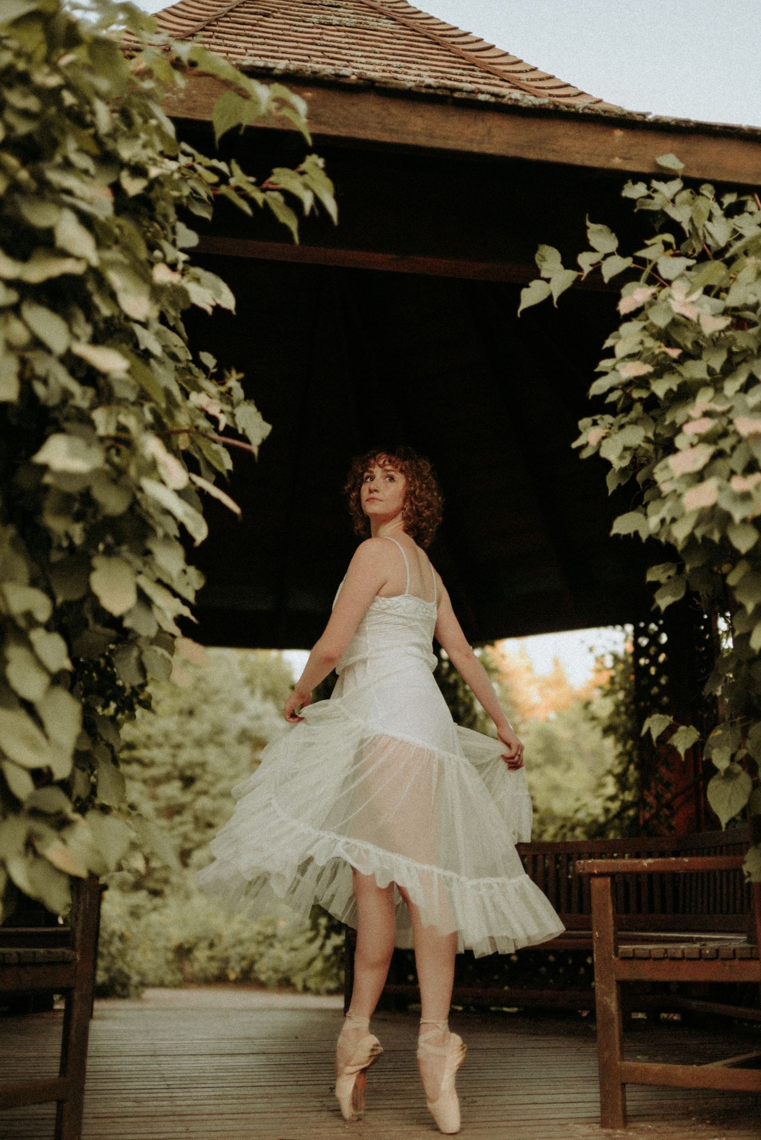 A dancer in a white tulle dress twirls gracefully on a wooden deck surrounded by lush green foliage.