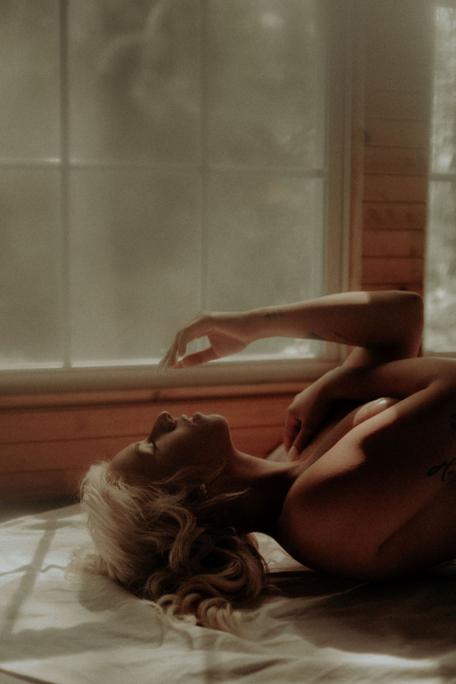 Artistic nude silhouette resting by window with wooden panels.