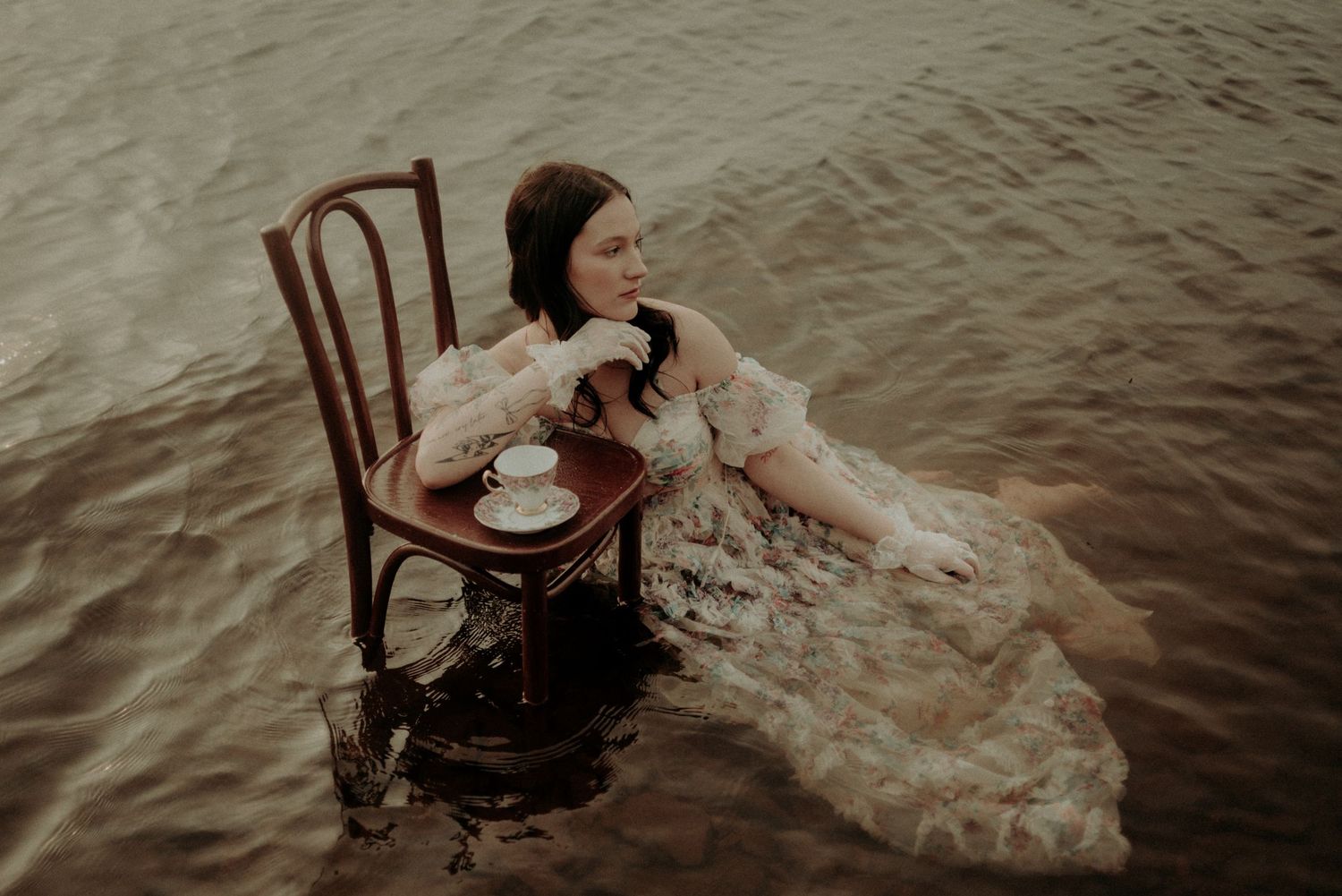 A vintage wooden chair sits in shallow water with a flowing dress creating an artistic scene.