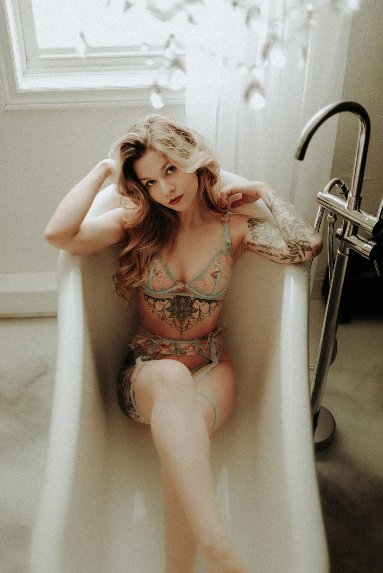 A moody boudoir portrait shows a woman with tattoos posing in lingerie in a white bathtub.