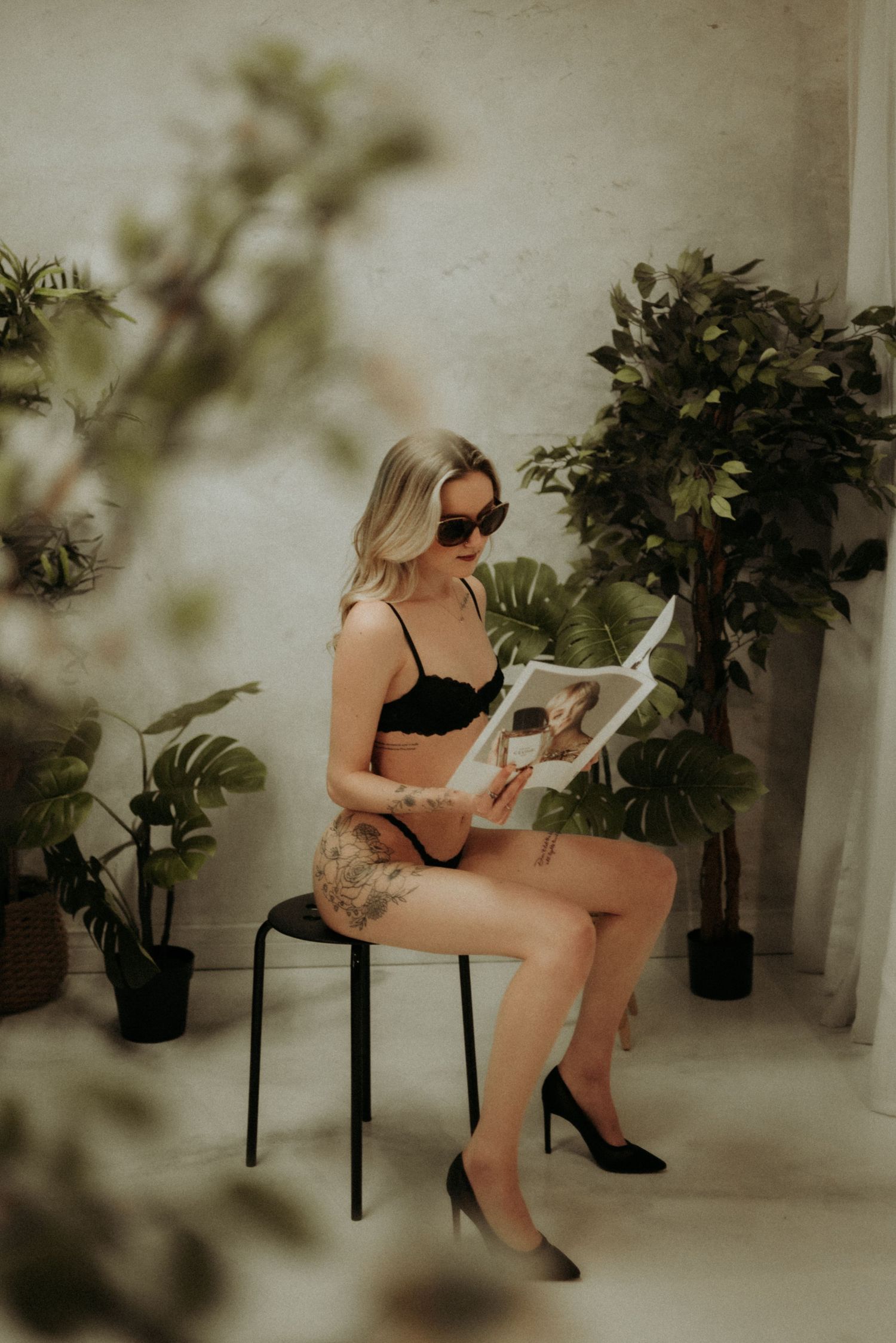 A woman in vintage-inspired lingerie reads a magazine while seated among potted plants in a dreamy boudoir setting.