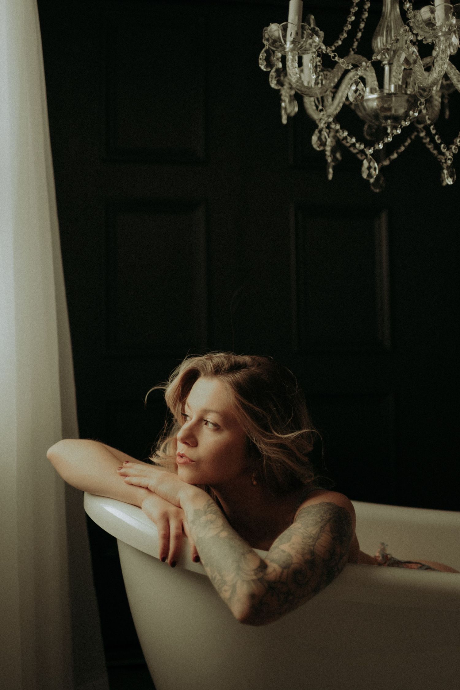 A serene artistic portrait of a figure resting in a clawfoot bathtub beneath a crystal chandelier.