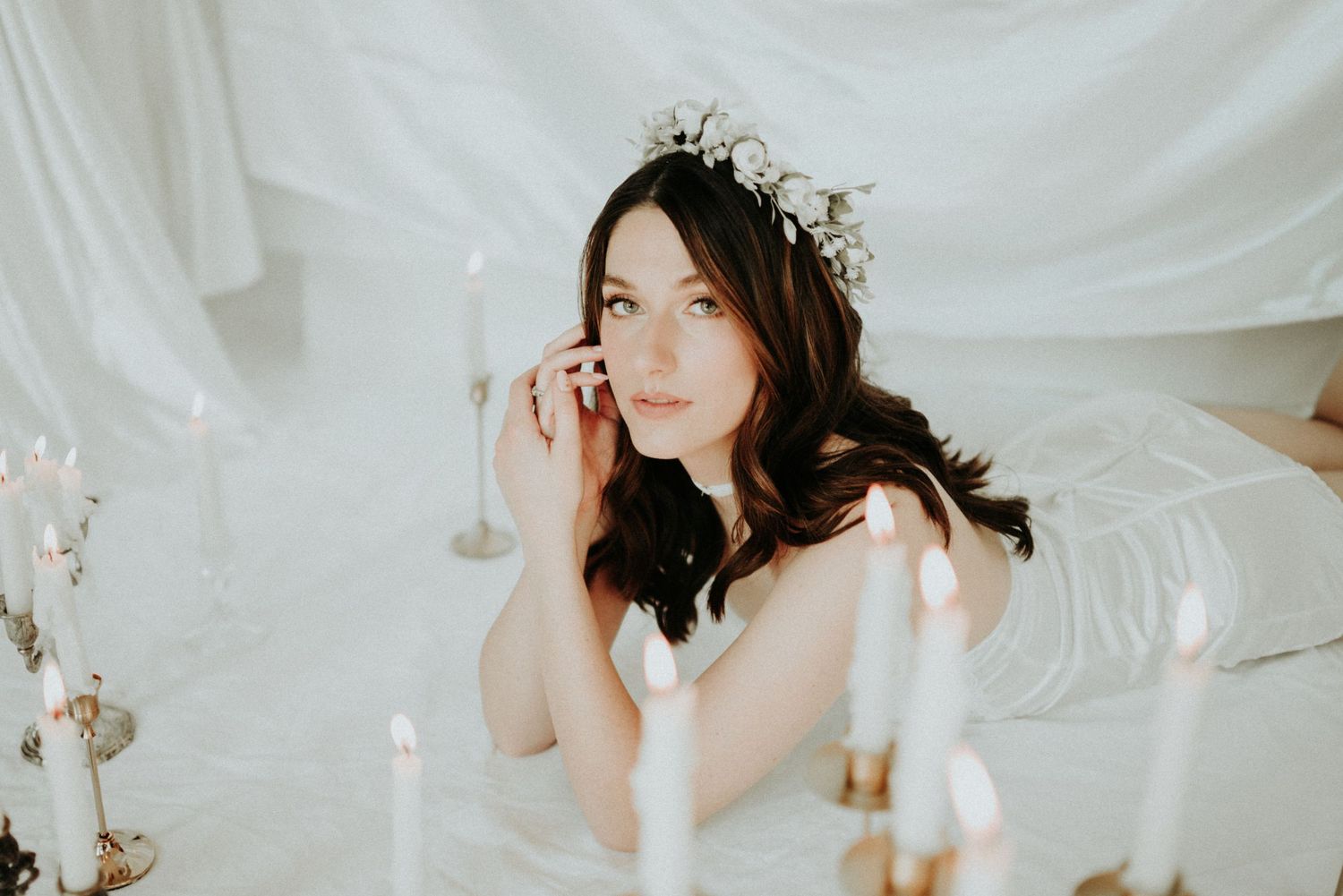 Bride wearing delicate floral crown sits at table with white backdrop and soft candlelight.