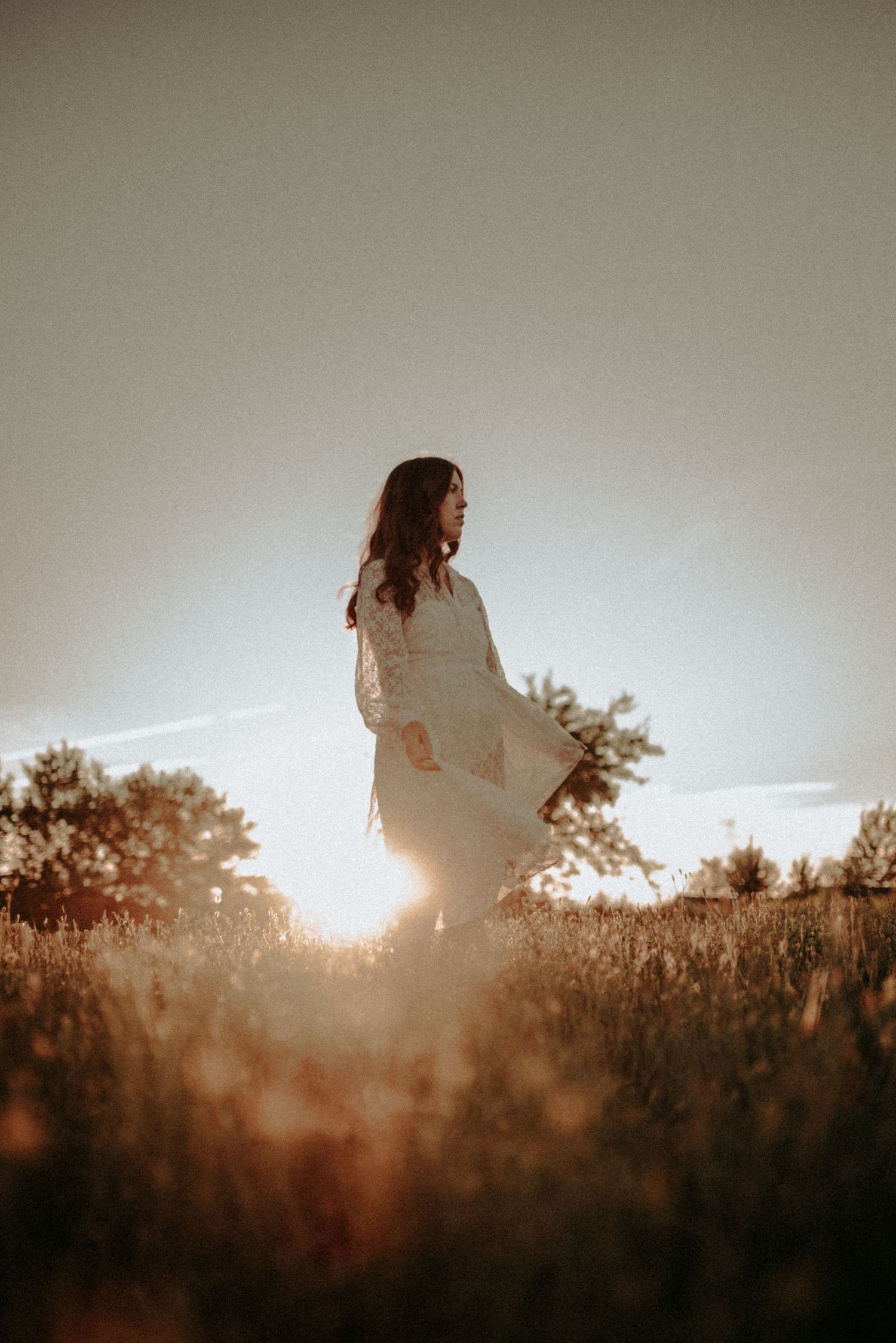 A silhouette stands in a golden field at sunset wearing a flowing white dress as sunlight creates a dreamy backlit effect.