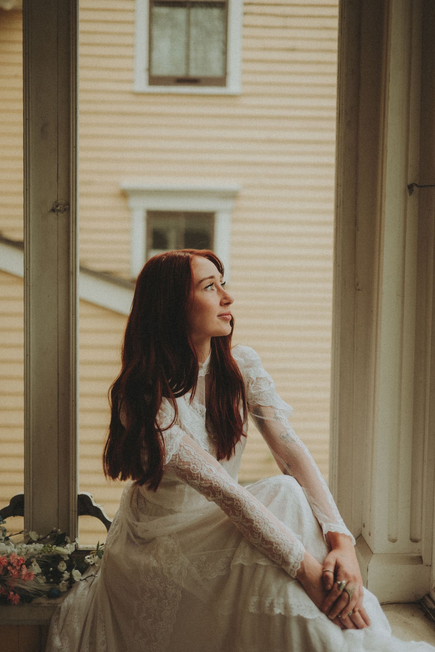 A dreamy vintage-style photo shows someone in a lace wedding dress sitting by a sunlit window with pale yellow siding outside.